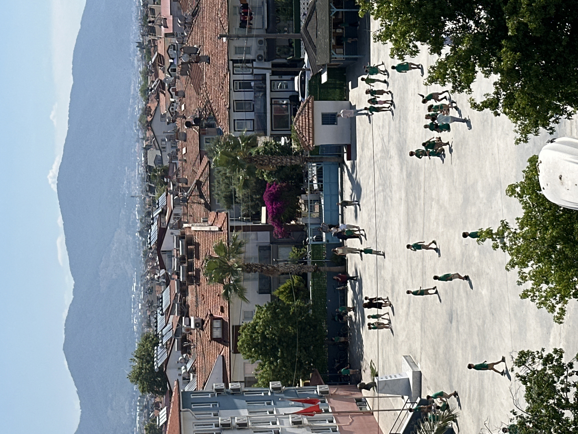 The image captures an elevated view of an outdoor scene, likely a schoolyard or public square, set against a backdrop of a town and distant mountains under a clear blue sky.
In the foreground and mid-ground, a group of approximately 20-30 individuals, mostly children or young adults, are spread across a light-colored paved area. They are predominantly dressed in green tops and dark bottoms, suggesting a uniform or specific attire for an event. Many are in various stages of movement, appearing to be performing a synchronized physical activity, drill, or parade. Their long shadows are cast on the ground, indicating bright sunlight, likely during the mid-morning or late afternoon. One adult, dressed in a lighter shirt, is seen observing or leading a smaller cluster of participants.
The paved area is bordered by various buildings. To the left and extending into the background are numerous multi-story structures featuring characteristic reddish-orange tiled roofs. Many of these rooftops are equipped with solar water heaters. Lush green trees, including palm trees and bougainvillea with purple flowers, are interspersed among the buildings, adding greenery to the urban landscape.
In the far background, a prominent mountain range rises against the sky, partially shrouded in a light haze, suggesting distance. The overall weather appears sunny and clear. The architectural style, vegetation, and mountainous terrain align with the provided context of Fethiye, Turkey. No discernible text is visible within the image.