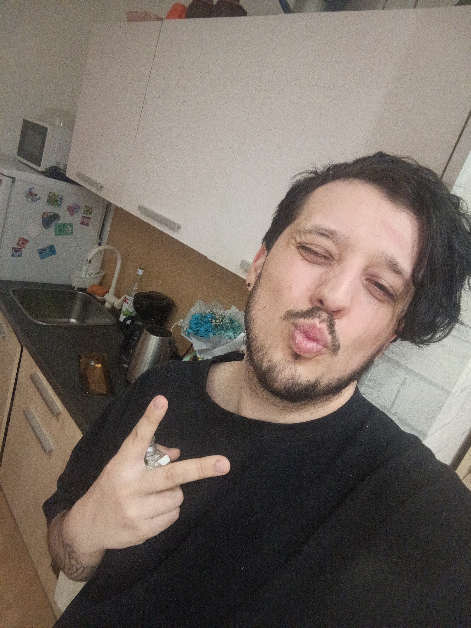 A man is taking a selfie in what appears to be a kitchen, likely in Penza, Russia, given the context. He is positioned in the foreground, facing the camera directly with a playful expression, winking with one eye and pursing his lips into a kissing face. He has dark, wavy hair, with part of it swept to his right side, and a beard and mustache. He is wearing a black, short-sleeved top, and a tattoo is visible on his left forearm. With his left hand, he makes a gesture similar to a "rock on" or peace sign, holding a small, metallic, pipe-like object between his index and middle fingers.
The background shows a home kitchen setting. To the man's left, there is a white refrigerator adorned with several colorful magnets. A white microwave oven sits atop the refrigerator. Adjacent to the refrigerator is a dark countertop with a built-in stainless steel sink and a white faucet. Various kitchen items are on the counter, including what looks like a black kettle or coffee maker, a small, dark brown rectangular object (possibly a phone or wallet), and a small bouquet of blue flowers wrapped in white paper. Above the counter, there are white kitchen cabinets with silver handles. The wall behind the man, to his right, features light-colored, possibly brick-patterned, tiling. The overall lighting suggests an indoor setting during the daytime. There is no visible text.