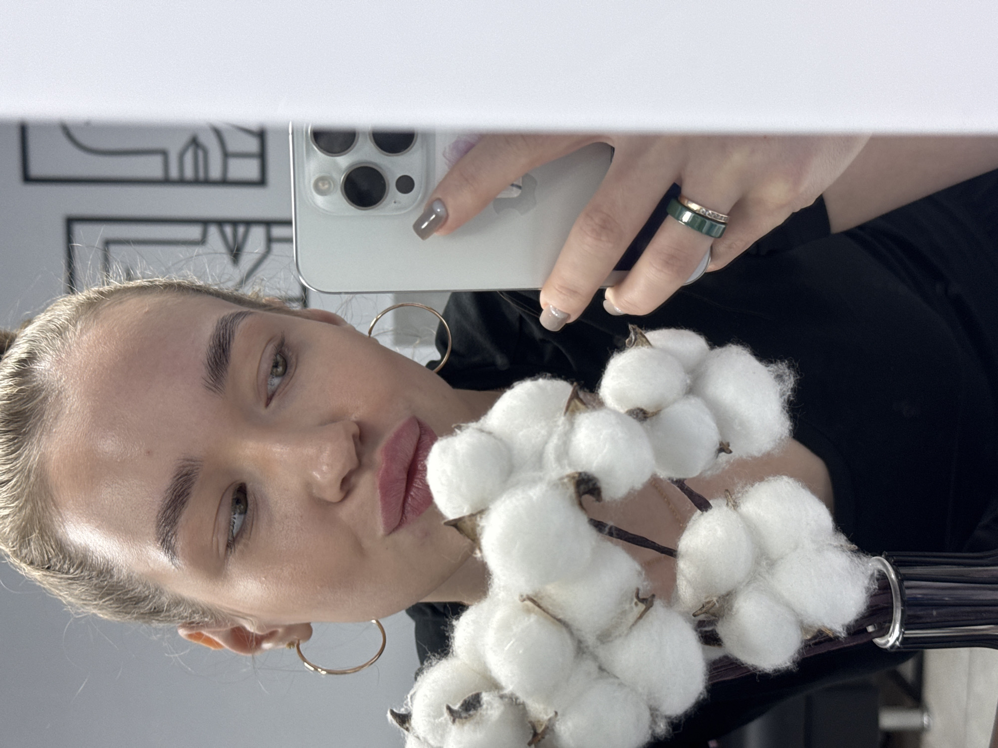 A young woman is taking a selfie in a mirror, holding a bouquet of cotton flowers. She has blonde hair pulled back, large gold hoop earrings, and wears a dark top. She is holding a white smartphone. On her finger, there is a green and gold ring. The reflection in the mirror shows a patterned design on a wall. The location is inferred to be Chaikovsky, Russia.