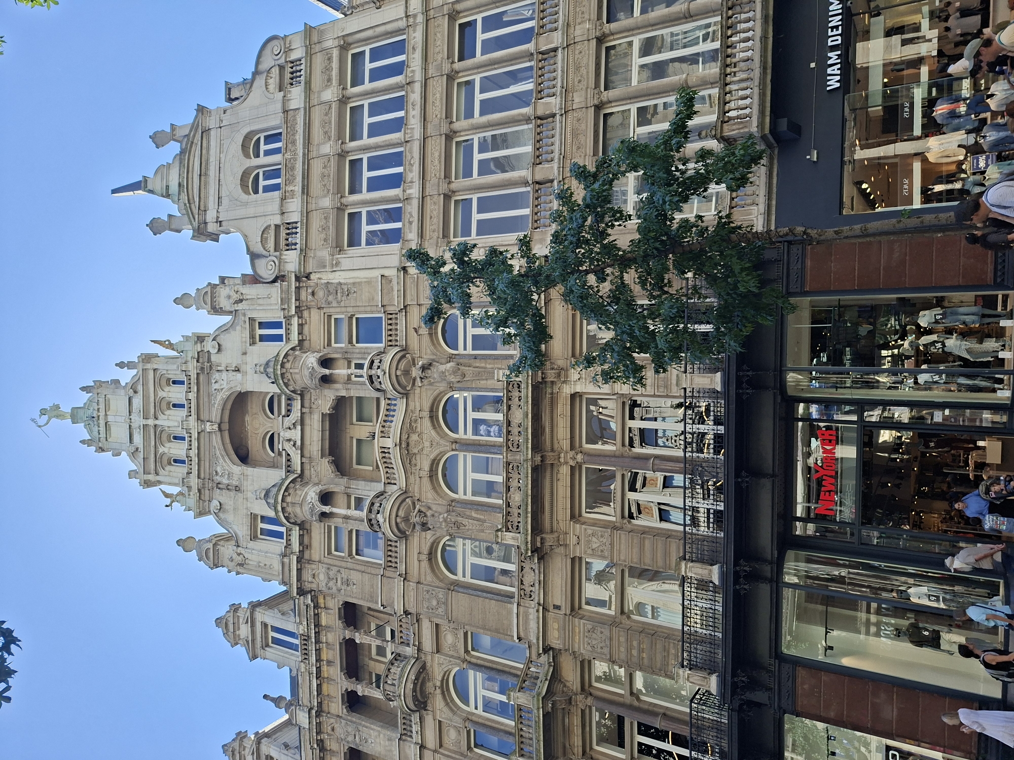 This is a low-angle, eye-level, full shot of a grand, ornate building on a city street in Antwerp, Belgium. The building's facade is richly decorated with sculptures, carvings, and arched windows, indicative of its historic architecture. A tree with dense green foliage stands in front of the building, partially obscuring the lower floors.
The ground level features storefronts. To the left, a "New Yorker" store is visible, with mannequins displayed in the window. To the right, a "WAM DENIM" store is also present, with a similar display of mannequins and clothing. The street is populated with a small number of pedestrians walking and browsing. The sky above is clear and blue, suggesting a sunny day. The image captures a bustling urban scene, highlighting the blend of historic architecture with modern retail.