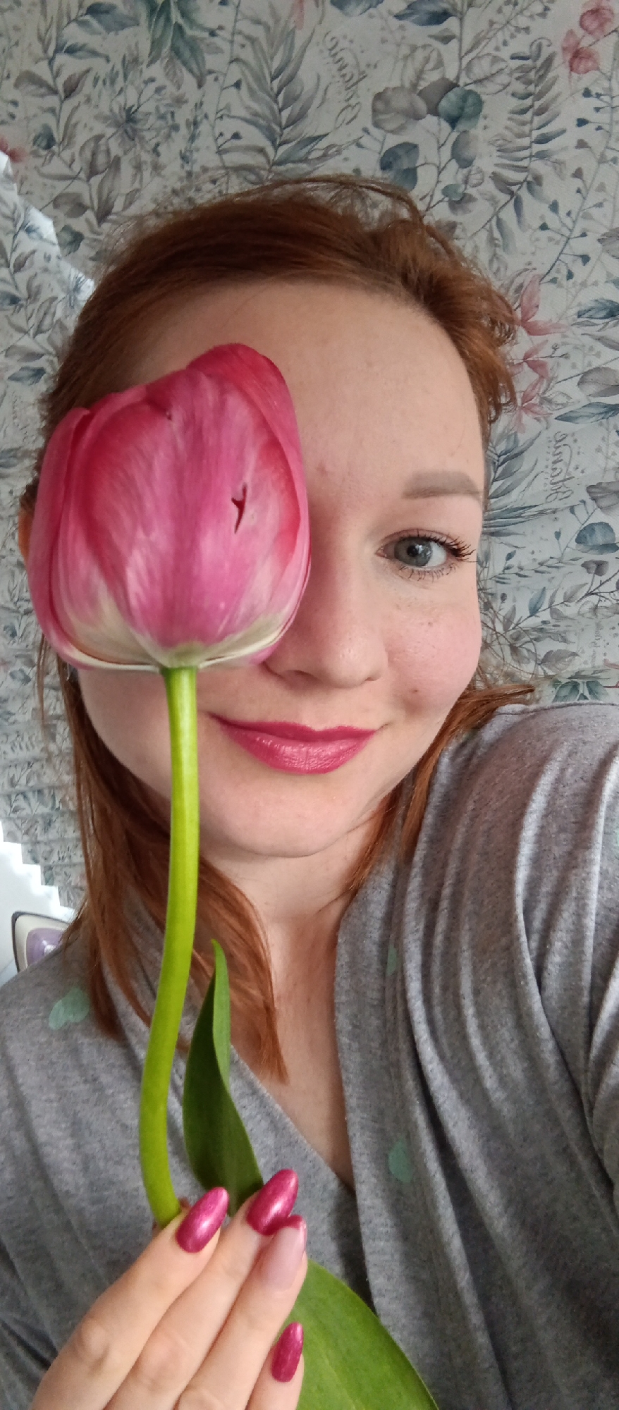 A person with red hair and pink lipstick holds a pink tulip in front of their face. The background is a floral patterned wallpaper. The location is Unknown, Unknown. The person's nails are painted pink. They are wearing a gray top. The person is smiling.