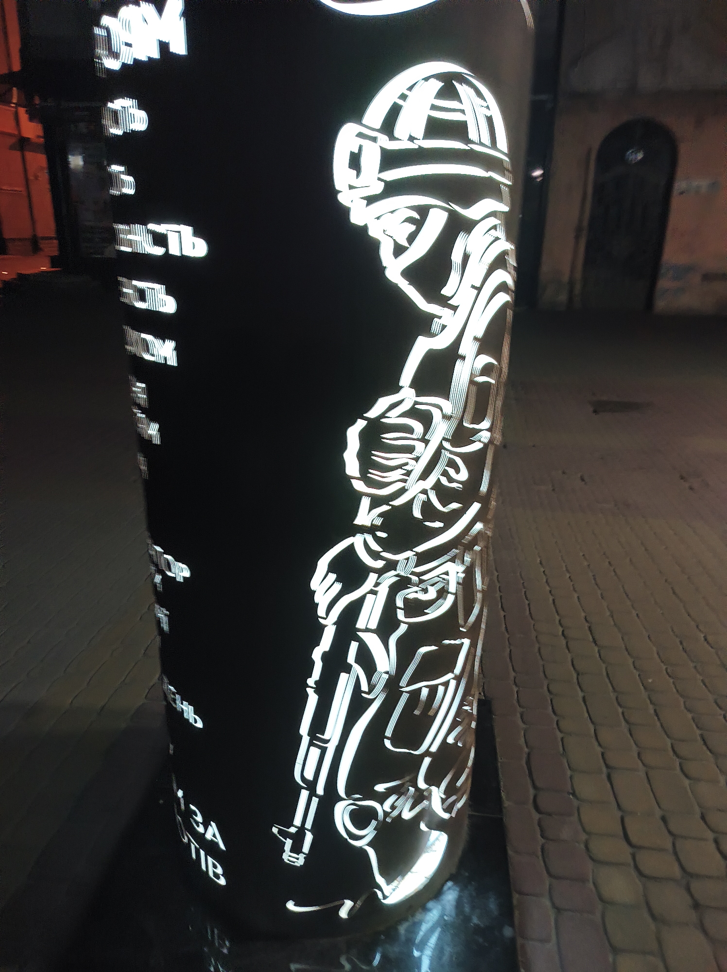 A nighttime photograph shows a bollard in Ternopil, Ukraine. The bollard is black with white illuminated outlines. On the right side of the bollard is an outline of a soldier with tactical gear and a rifle. On the left side are vertical illuminated white Ukrainian words. The bollard sits on a cobblestone walkway. A building is visible in the background with a dark arched door.