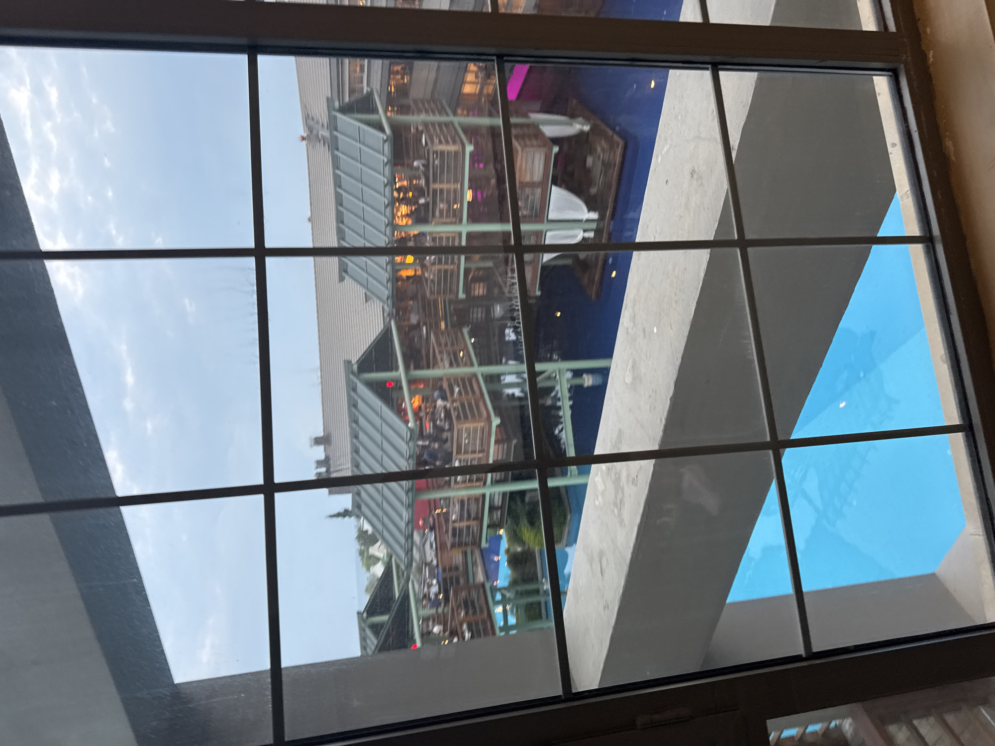 The image, taken from a window with a grid pattern, shows an outdoor dining complex built over a body of water, likely a swimming pool. The structures consist of multi-story wooden buildings with green roofs, resembling elevated restaurants or bars. People are visible on the balconies and upper levels of these structures, suggesting they are occupied. Some areas of the buildings are illuminated with warm, yellow lights. Below the structures, the water appears a vibrant blue, with some floating elements that might be seating areas or decorative features. The overall environment suggests a resort or entertainment area. The location is identified as Aksu, Turkey, a region known for its tourism and coastal resorts. The weather appears to be overcast with some clouds visible in the sky.