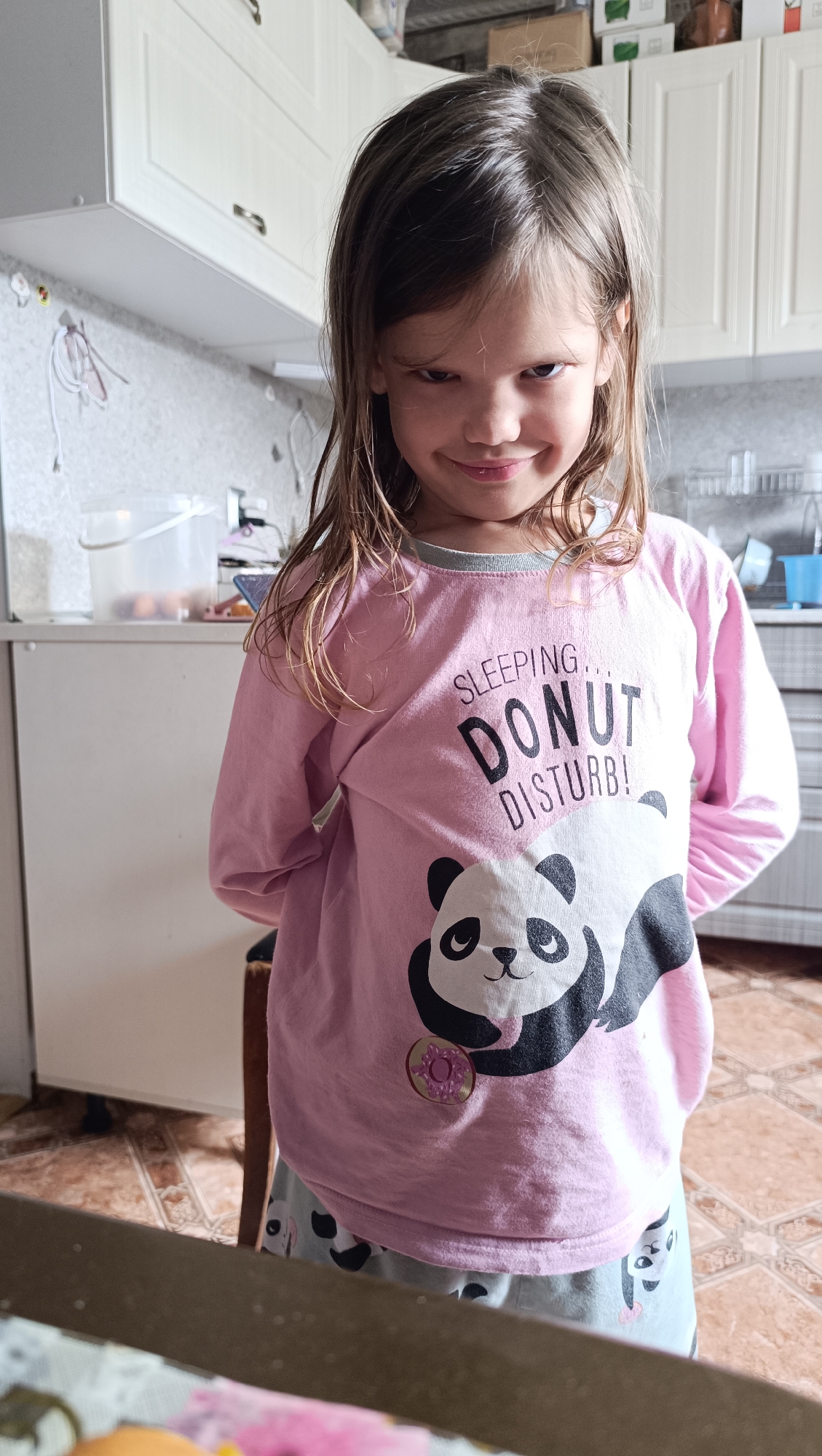 A young girl with brown hair pulled back stands in a kitchen wearing a pink long-sleeved shirt with a panda and donut design and grey pants with panda faces. She is looking directly at the camera with a sly smile and her hands behind her back. The kitchen has white cabinets, a tiled floor, and a countertop in the foreground. The image appears to be taken in Karsha, Russia.