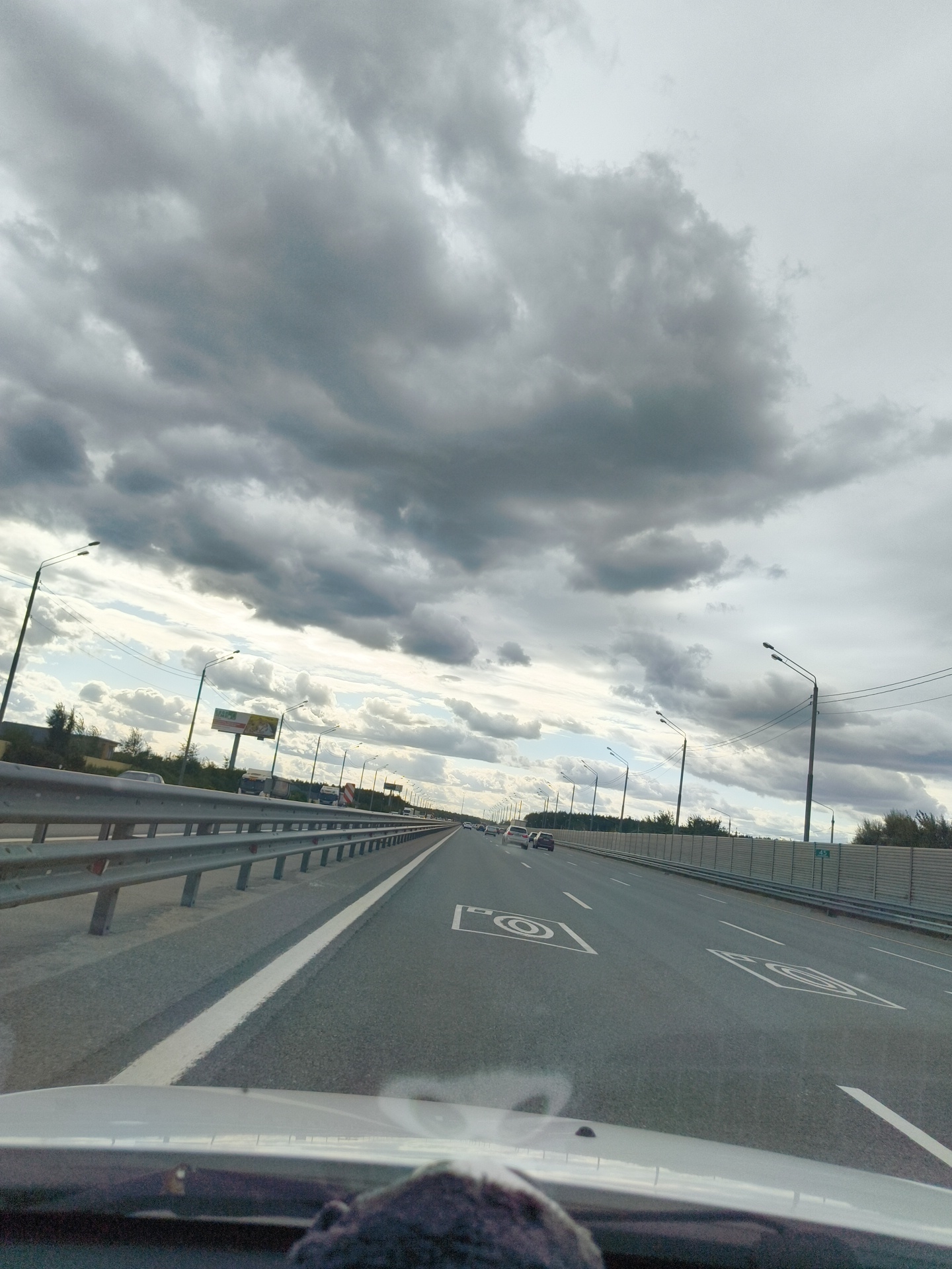 The image depicts a view from inside a vehicle traveling on a multi-lane highway. The sky is overcast with dramatic, dark clouds. The highway has distinct lane markings and speed limit indicators (indicated by a camera symbol and the number '45') painted on the asphalt. Guardrails line the side of the road, and streetlights are visible at intervals. Vegetation and what appear to be industrial structures or sound barriers are present on the right side of the highway. Several vehicles are traveling in the same direction. The scene suggests movement along a transportation artery, possibly on the outskirts of Zdorov'ye, Russia, under a stormy sky.