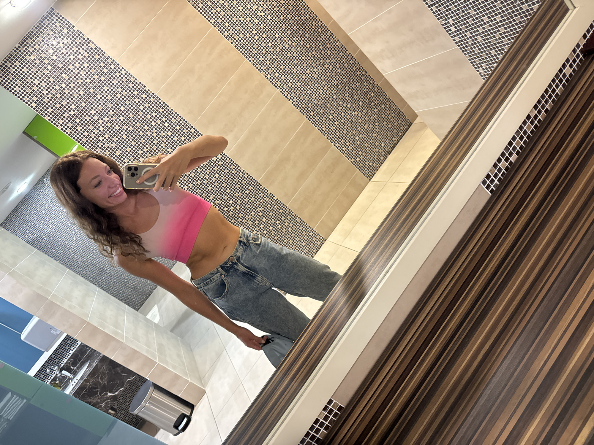 The image shows a woman taking a selfie in a restroom. The woman has long curly brown hair, wearing a pink and white top and blue jeans. She is holding a cell phone in her right hand and smiling towards the camera. The restroom has tiled walls with horizontal bands of beige and dark mosaic tiles. The vanity area has a stainless steel trash can and a black countertop. There is a mirror on the right side of the frame with a striped wood-grain border. The location is presumably in Đà Nẵng, Vietnam, but there is no definitive evidence to support this claim in the image itself.