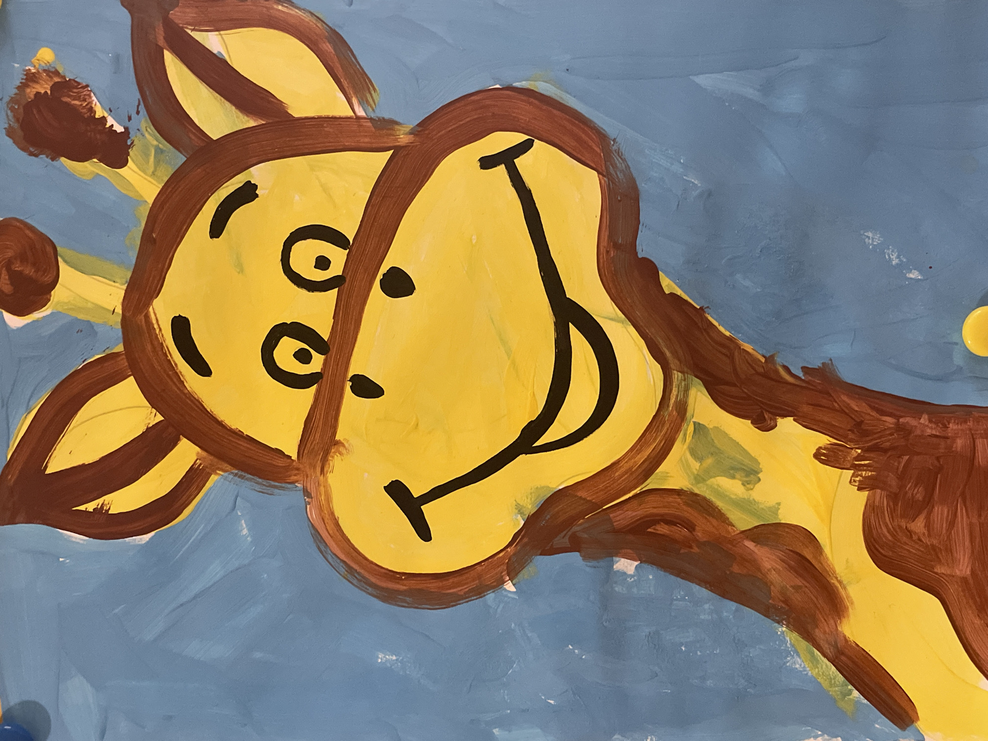 The image is a close-up, eye-level, vertical shot of a painting of a cartoon giraffe's head and neck against a blue background. The giraffe is bright yellow with brown markings around its eyes, ears, horns, and neck. It has large, black, circular eyes and a wide, black, curved smile. The painting style is simple and appears to be done by a child, with visible brushstrokes and some smudges. The background is a solid, light blue with visible texture from the paint. There are two small yellow circular objects visible at the bottom edge of the frame, appearing to be pushpins or tacks. No specific location in the city of Unknown, Unknown can be determined from the image.