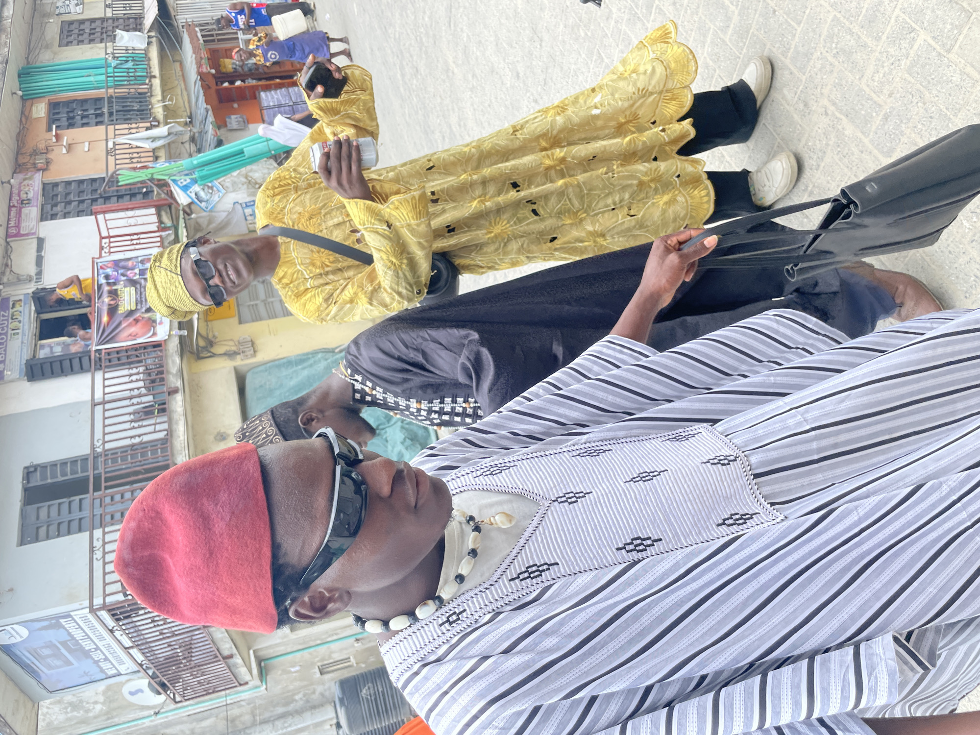 Three men stand in a street in Ado, Nigeria. The closest man wears a red cap, sunglasses, a striped shirt, and a necklace. Behind him, a man in a black robe is partially visible. The third man wears a yellow robe with a matching head covering and sunglasses. He is holding a white object in his hand. A black bag is also visible. The background includes buildings and storefronts.