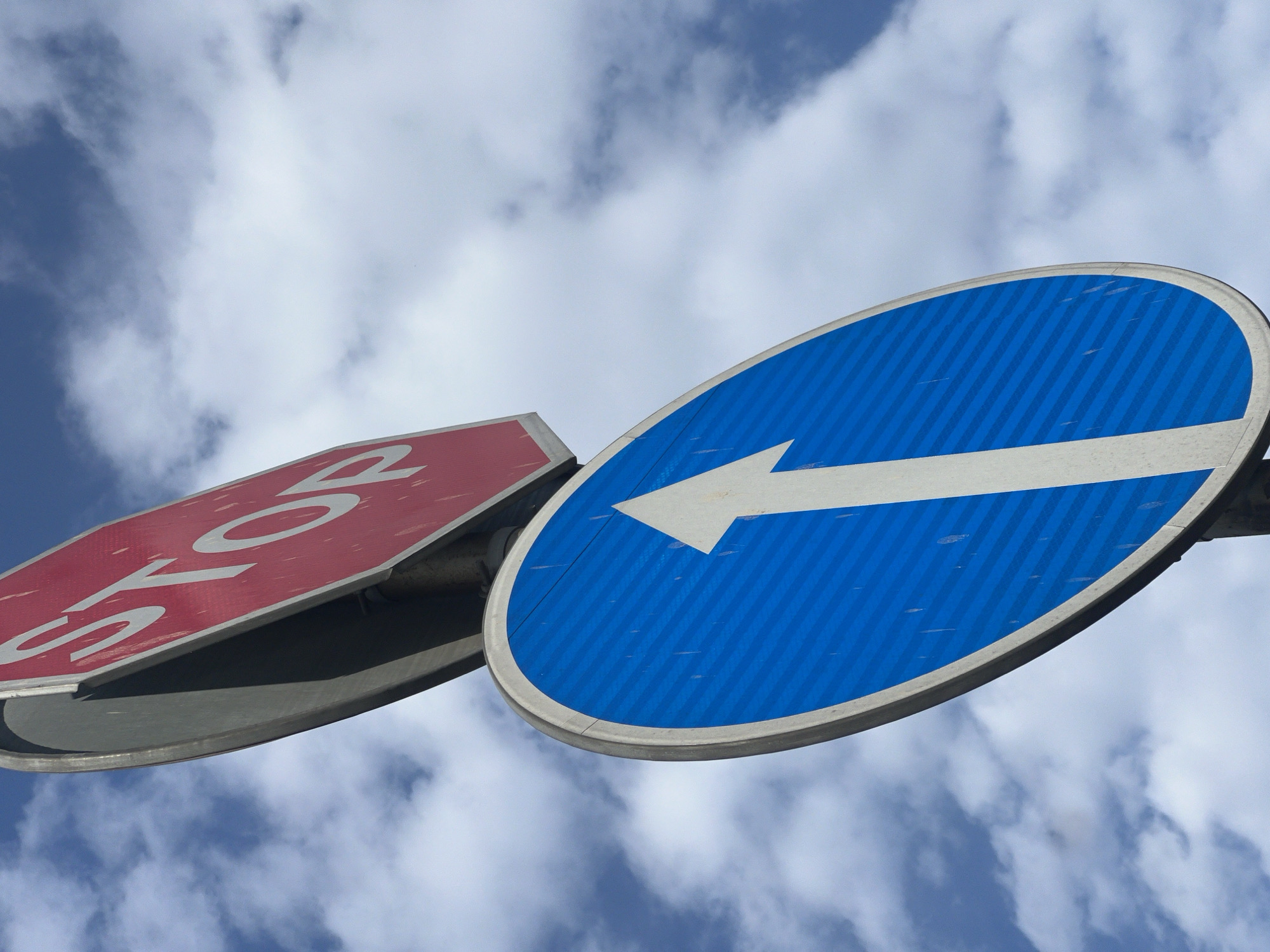 The image displays a low-angle view of two road signs against a bright, cloudy sky in Yerevan, Armenia. In the foreground, on the left, is a red octagonal sign. The visible white text on this sign clearly spells "STOP," indicating a mandatory stop. The sign appears somewhat weathered or dusty, with its underside also visible, suggesting it's mounted on a pole.
Adjacent to and slightly overlapping the stop sign, on the right, is a blue circular sign. This sign features a prominent white arrow pointing horizontally to the left, typically signifying a mandatory turn left or an instruction to keep left. The blue background of this sign has a subtle striped or textured pattern, and like the stop sign, it shows minor surface imperfections.
Both signs are angled, viewed from below, and positioned against a backdrop of a blue sky dotted with fluffy white and light grey clouds. The lighting suggests it is daytime. The scene focuses directly on these two informational traffic signs, essential for guiding drivers and pedestrians.