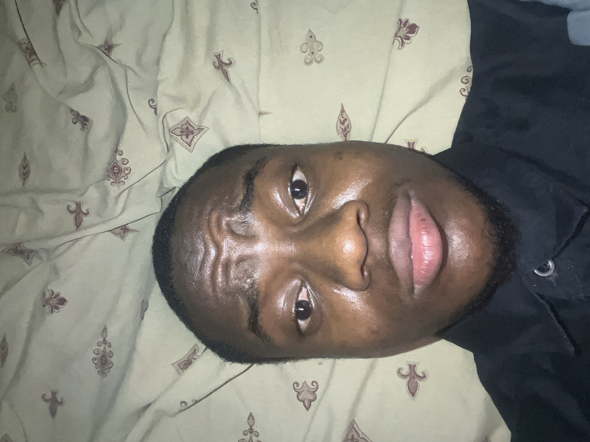 A person lies supine on a bed covered with patterned beige linens. The person has dark skin, short hair, and a beard. They are wearing a dark-colored shirt. The location is not discernible from the image. Therefore, it is impossible to determine if the image was taken in Minna, Nigeria.
