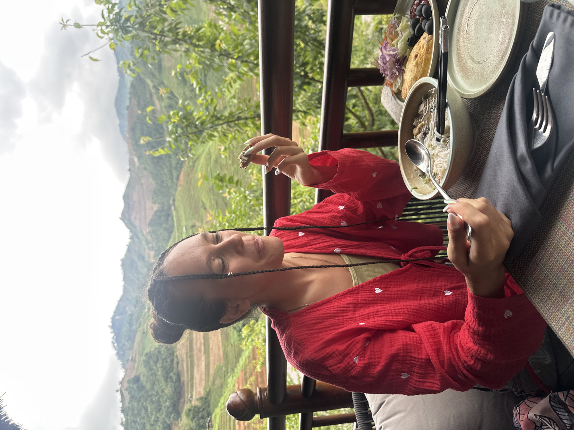 This image captures a woman seated at an outdoor table with a mountainous, terraced landscape view in Púng Luông, Vietnam, under an overcast sky. The woman has dark skin and her braided hair is styled in a bun. She wears a red, long-sleeved, textured blouse adorned with small white embroidered hearts, layered over a light green tank top. Her left hand, with manicured nails and rings, holds a small, dark piece of food near her mouth, while her right hand holds a spoon in a bowl of noodles with various ingredients, including mushrooms and what appears to be a clear broth. Chopsticks are also resting in the bowl. On the table, to the right of the noodle bowl, an empty round plate is present, and another plate holds a meal consisting of a golden-brown pastry or fried item, several black olives, and a salad of shredded purple cabbage and other greens. The table surface is a patterned, light-brown textured placemat. A dark grey napkin, holding a fork and knife, is positioned on the table. A dark wooden railing with a decorative post runs in front of her, separating the dining area from the expansive natural environment. Beyond the railing, vibrant green foliage and trees are visible, giving way to rolling hills covered in intricate agricultural terraces, likely rice paddies, extending under the cloudy sky.