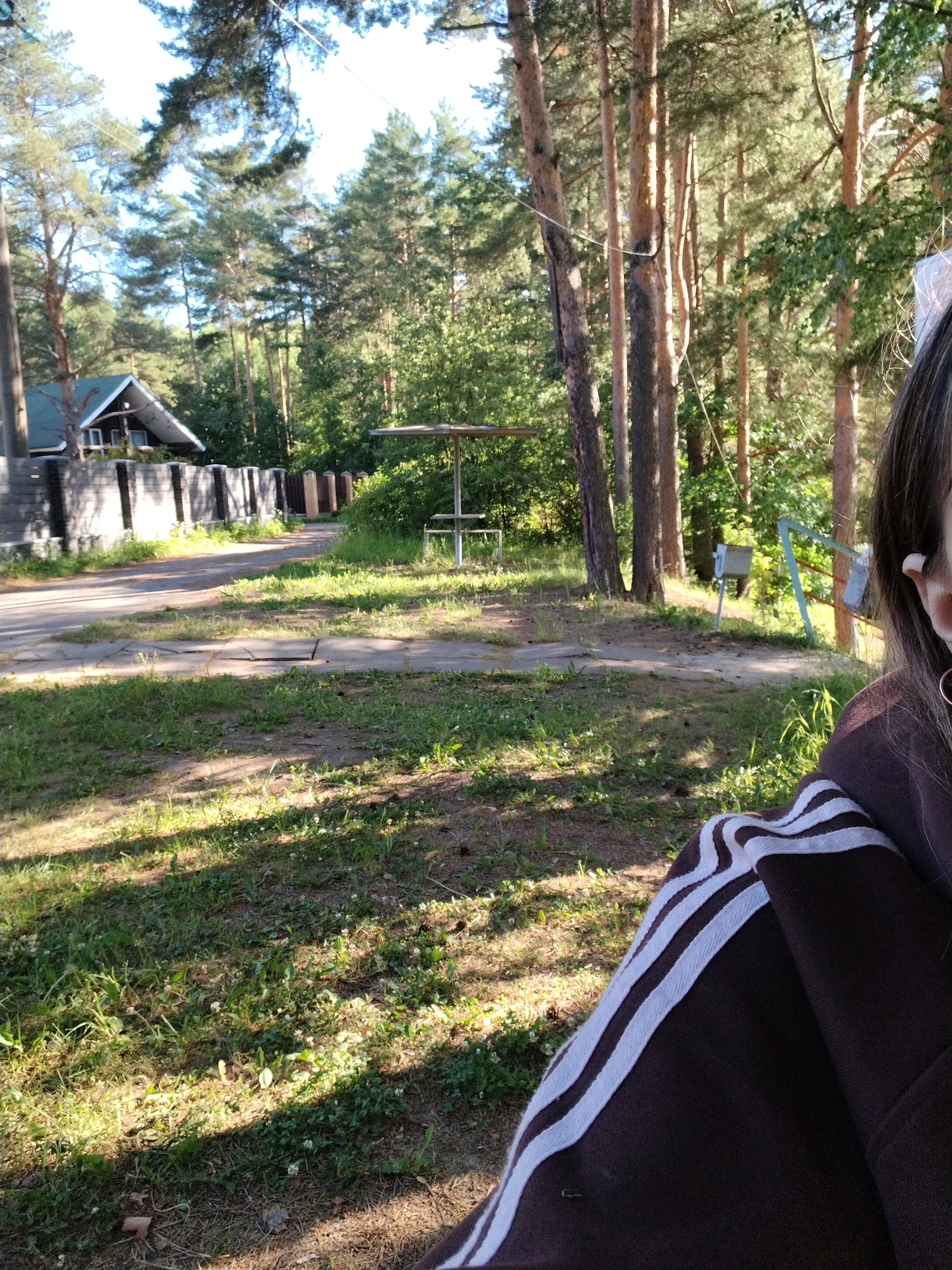 The image depicts an outdoor scene, likely in a suburban or rural area, with elements suggesting a location in Stroitel', Russia. In the foreground, a portion of a person wearing a dark brown hoodie with white stripes on the sleeve is visible, occupying the lower right quadrant of the frame. The person is looking towards the left, with their right ear and part of their face visible.
The background reveals a tree-lined area with tall, slender pine trees dominating the landscape. Sunlight filters through the trees, casting dappled shadows on the grassy ground. A paved road or pathway runs diagonally from the left towards the center, bordered by grass and some scattered debris. To the left of the road, a dark fence runs along the property line, with a portion of a house visible behind it. The house has a dark roof and windows. Further into the background, a small, open-sided shelter structure with a table and benches is visible, along with what appears to be a swing set with a metal frame. The overall environment suggests a park or recreational area adjacent to a residential property. The sky is clear blue with sunlight indicating a daytime scene.