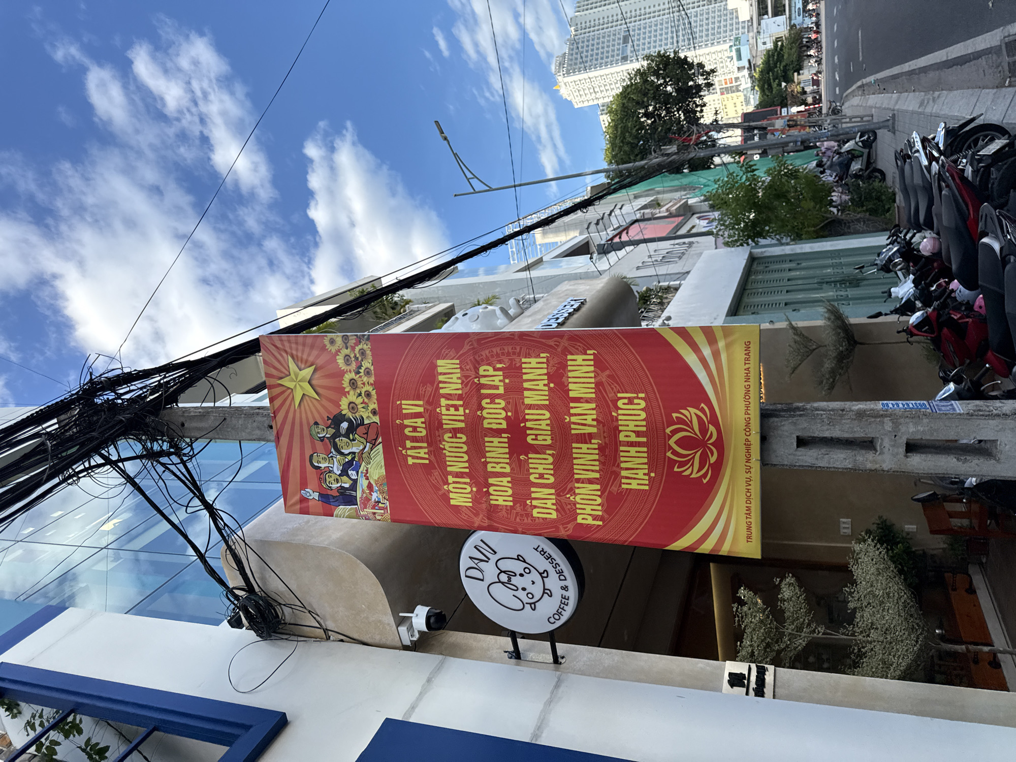 This is a medium-shot, eye-level, outdoor, daytime, vertical, color photograph taken on a street in Nha Trang, Vietnam. A red banner with yellow text and imagery hangs from a utility pole on the left side of the frame, advertising "TẤT CẢ VÌ MỘT NƯỚC VIỆT NAM HÒA BÌNH, ĐỘC LẬP, DÂN CHỦ, GIÀU MẠNH, PHỒN VINH, VĂN MINH, HẠNH PHÚC!". Beneath the banner, a circular white sign with a cartoon image of a creature and text reads "DANI COFFEE & DESSERT". The background consists of a modern building with a blue-trimmed window on the left and a glass facade reflecting the sky. A street runs along the right side of the frame, lined with parked motorcycles and a few pedestrians. In the distance, tall buildings and a clear blue sky with scattered clouds are visible. The scene depicts a commercial street in Nha Trang, with a focus on local businesses and a nationalistic message displayed on the banner.