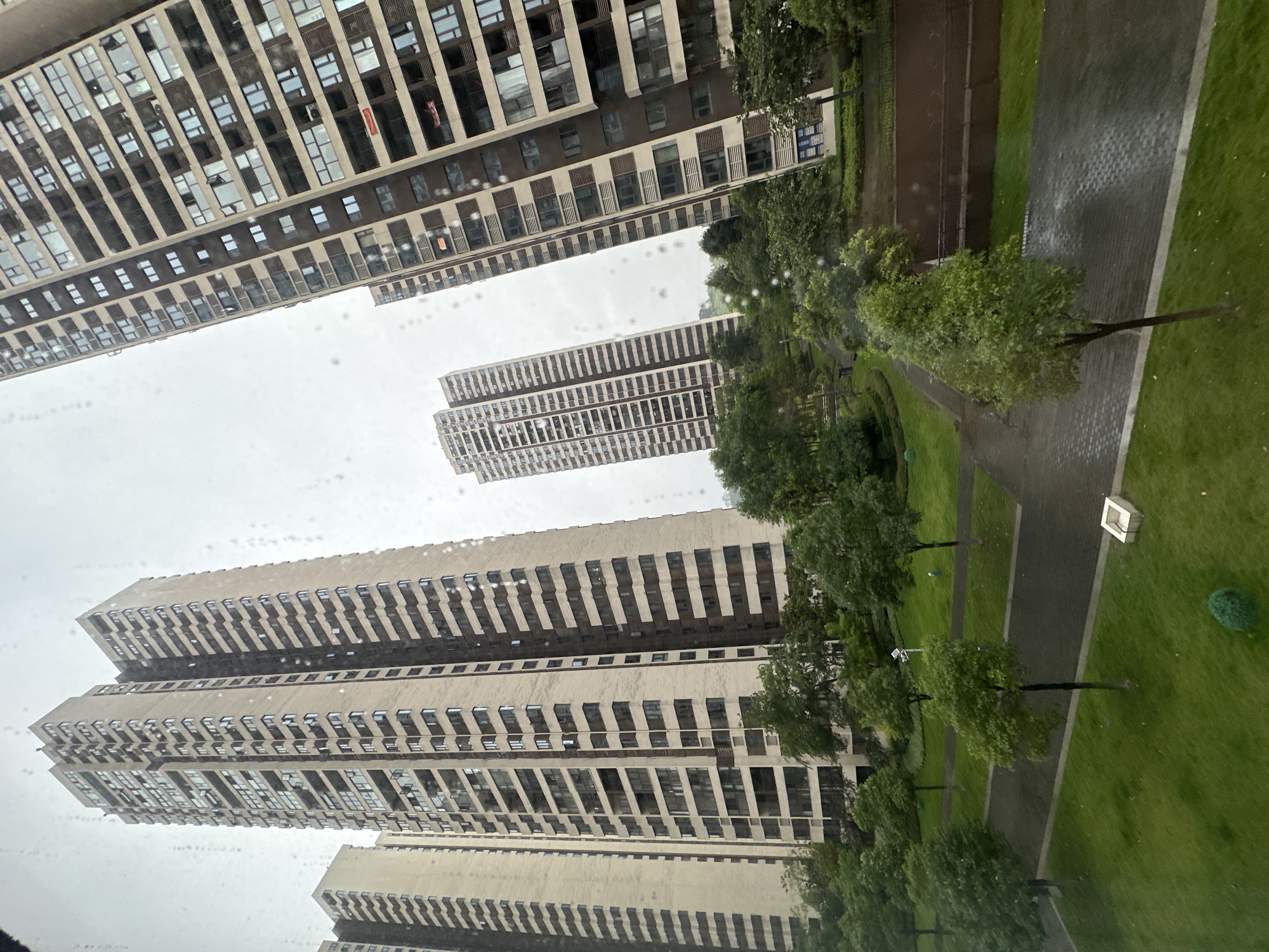This image captures a dense residential complex within the Chongqing High-tech Zone, China, on an overcast day, observed through a window speckled with water droplets, suggesting recent rain. The scene is dominated by multiple towering apartment buildings with extensive green spaces below.
On the left, two tall, light beige-colored residential towers rise prominently, featuring numerous stacked balconies and windows. Further left and slightly behind, another similar building is partially visible. In the mid-distance, two more high-rise buildings, slightly lighter in hue, stand against the grey sky. To the right, a large, dark brown residential building with distinctive broad horizontal bands and numerous windows, some open, occupies a significant portion of the frame. Several small, red banners or signs, possibly indicating "for rent" or "for sale," are affixed to balconies on the rightmost building.
The ground level is meticulously landscaped with lush green lawns and a variety of trees, mostly deciduous, forming an urban park-like environment. Paved pathways, some with a patterned texture, curve through the grassy areas, appearing wet in sections from recent precipitation. Visible elements within the green space include a small, white square planter or decorative object in the bottom right, a spherical green shrub near the bottom center, and a light pole in the mid-ground. No individuals or active vehicles are present, indicating a quiet moment in the complex.