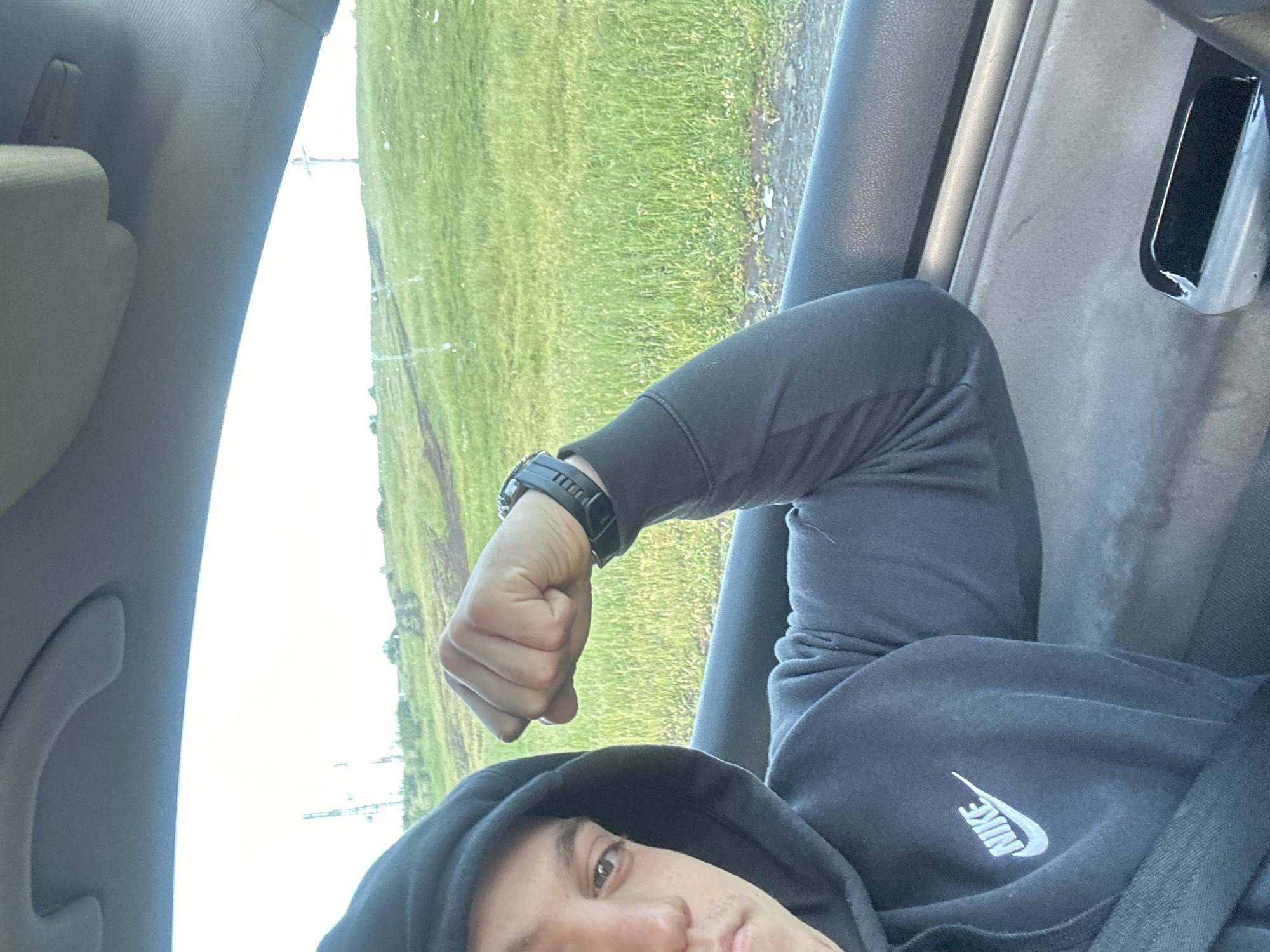 A man is inside a car, possibly in Burgas, Bulgaria, based on the landscape visible through the car window. The landscape consists of green fields, likely located on the outskirts of the city or a rural area near Burgas. He is wearing a dark hooded sweatshirt with a Nike logo. He is flexing his arm and has a black watch on his wrist. The interior of the car is visible, including the car door, paneling, and headliner. The image appears to have been taken from a low angle, potentially from a smartphone.