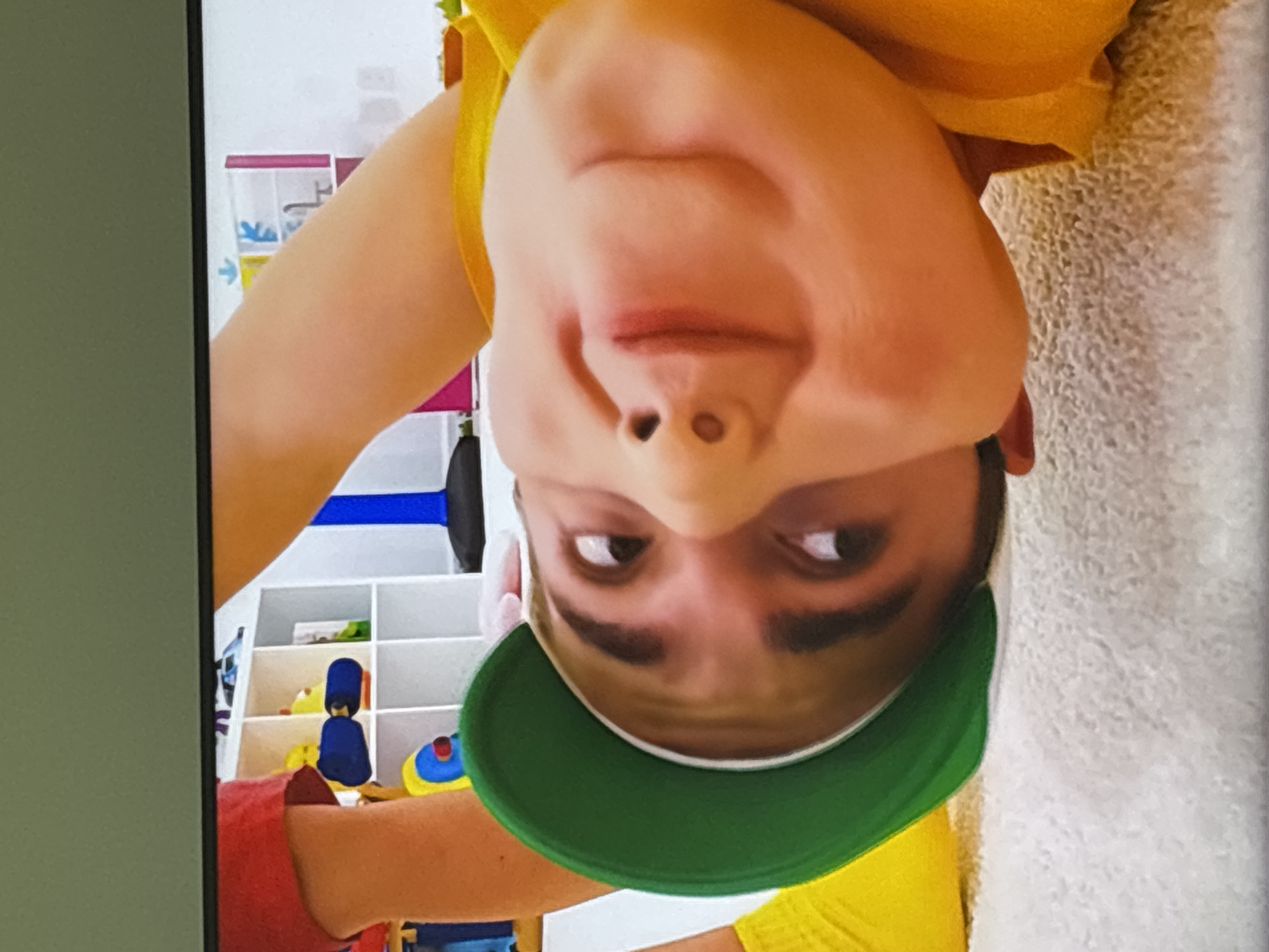 A close-up, inverted, low-angle, medium shot captures a young child lying on a textured, light-colored surface, possibly a rug. The child is wearing a bright yellow t-shirt and a green cap, which is positioned upside down on their head. Their face is the primary focus, with the eyes looking upwards and to the left, and the mouth slightly open, forming a subtle smile. The background is blurred but suggests an indoor setting, with shelves containing various items, including what appear to be toys. The overall lighting is bright, highlighting the child's features. The image does not provide any information to determine the location within Dubai, United Arab Emirates.
