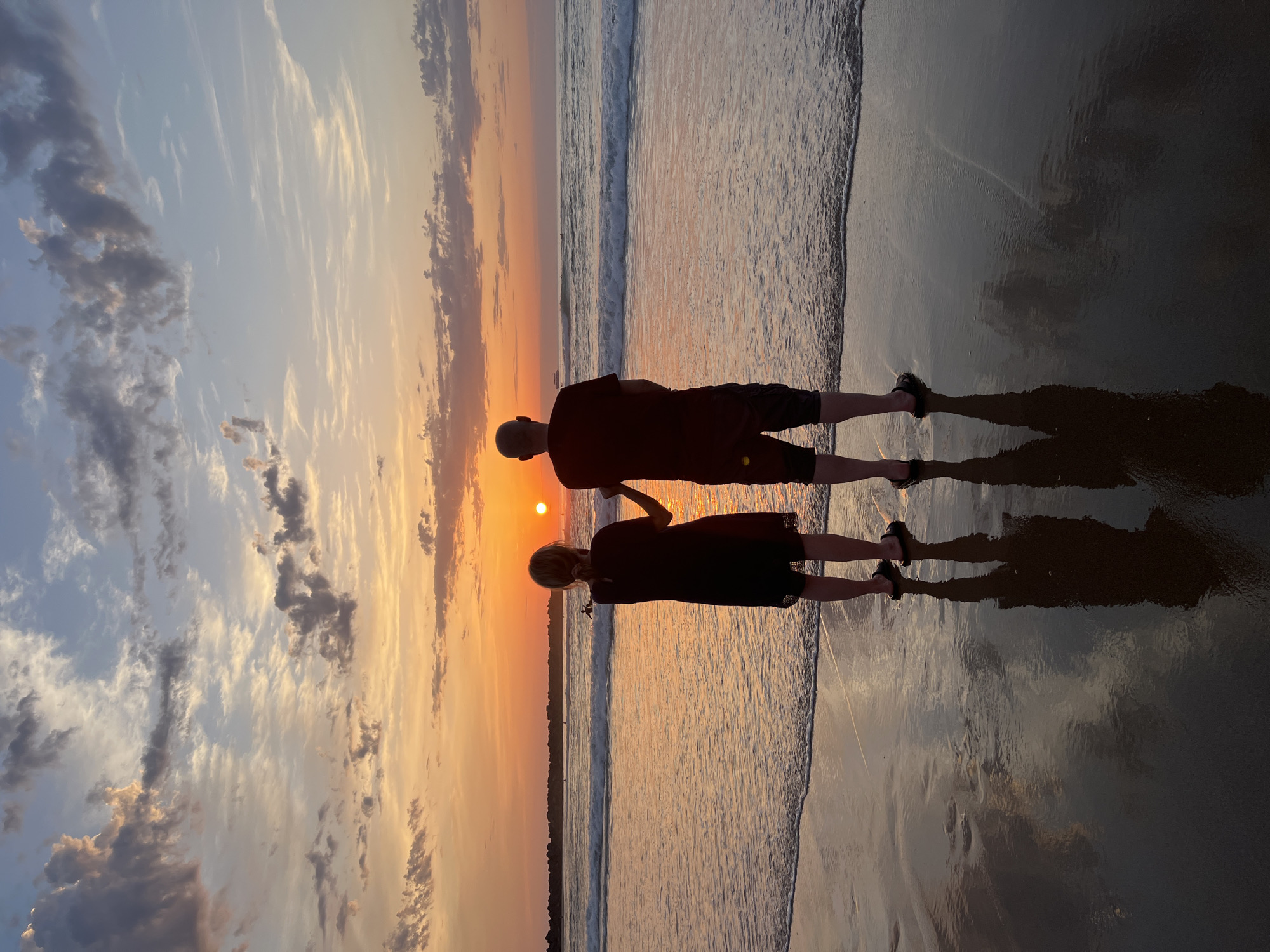 Two individuals, a woman and a man, stand on a wet sand beach, facing the ocean and a setting sun. The woman, on the left, wears a dark, knee-length dress and sandals. The man, on the right, wears a dark t-shirt and shorts, also in sandals. They are positioned close together, with the woman's left hand appearing to touch the man's right arm or hand. The sun, a bright orb, is visible low on the horizon, casting a warm orange glow across the sky and reflecting on the water's surface. Gentle waves roll towards the shore, creating a foamy edge on the wet sand where the couple stands. The sky is a gradient of soft blues, oranges, and pinks, with scattered clouds tinged by the sunset. The reflections of the couple and the sky are clearly visible on the wet sand. In the background, the ocean stretches to the horizon, with a few distant figures visible in the water, possibly surfers. The scene suggests a peaceful moment of observing the sunset. The location is La Concha Beach in San Sebastián, Spain, a well-known urban beach renowned for its crescent shape and scenic views, particularly at sunset.