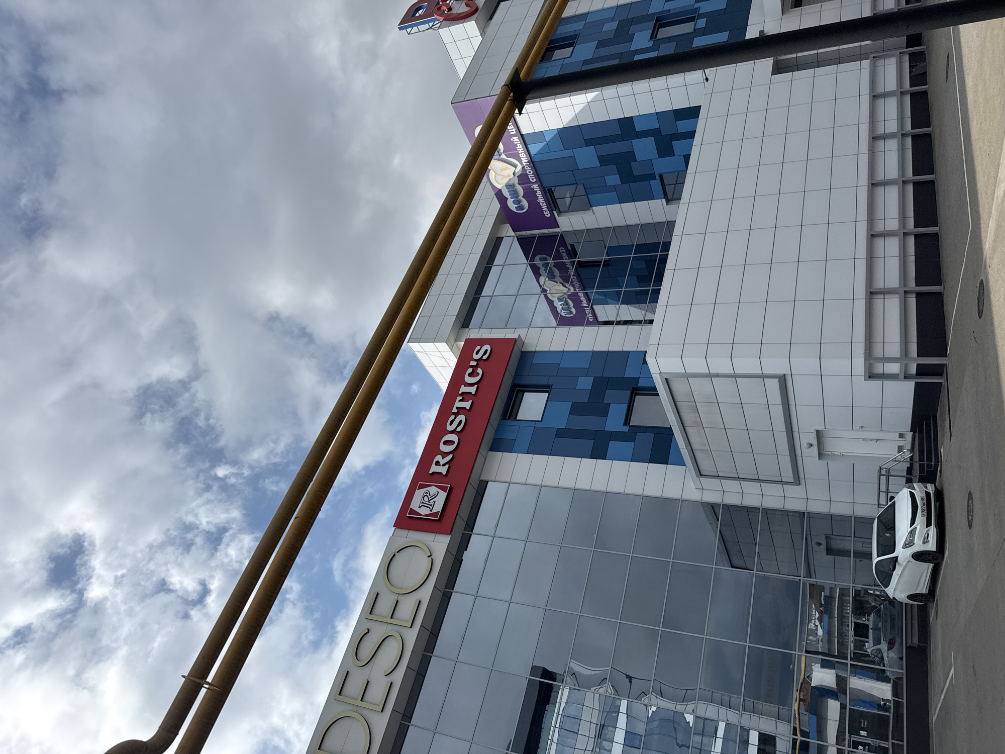 The image shows the exterior of a modern building with a white tiled facade and blue glass windows. Two large, rusty, cylindrical structures run diagonally across the frame from the bottom left. A red sign with white lettering reads "ROSTIC'S" and features a red "R" logo. Another sign with gold lettering spells out "DESEO" vertically. The building has multiple floors with a blue and white patterned design on some sections. In the lower right, a white car is parked on an asphalt surface marked with white lines. The sky above is cloudy with patches of blue peeking through. The scene appears to be outdoors on a cloudy day. The location is identified as Sochi, Russia, and the image depicts a commercial or retail building. There are no people visible in the image.