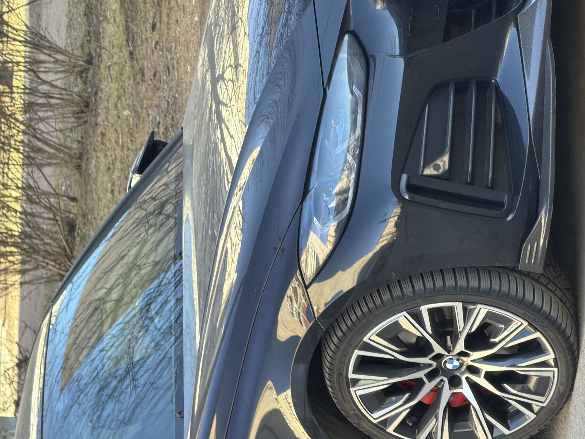 The image shows a close-up of a black BMW car. The focus is on the front driver's side, highlighting the headlight, part of the front grill, the wheel, and a small portion of the car's body. The wheel has a distinctive design with a BMW logo in the center, and red brake calipers are visible. The background is blurred, suggesting an outdoor environment with some trees or vegetation. Given the location in Saint Petersburg, Russia, the scene likely depicts a parked car in a residential or urban area of the city. The lighting indicates it's daytime, with sunlight reflecting off the car's surface.