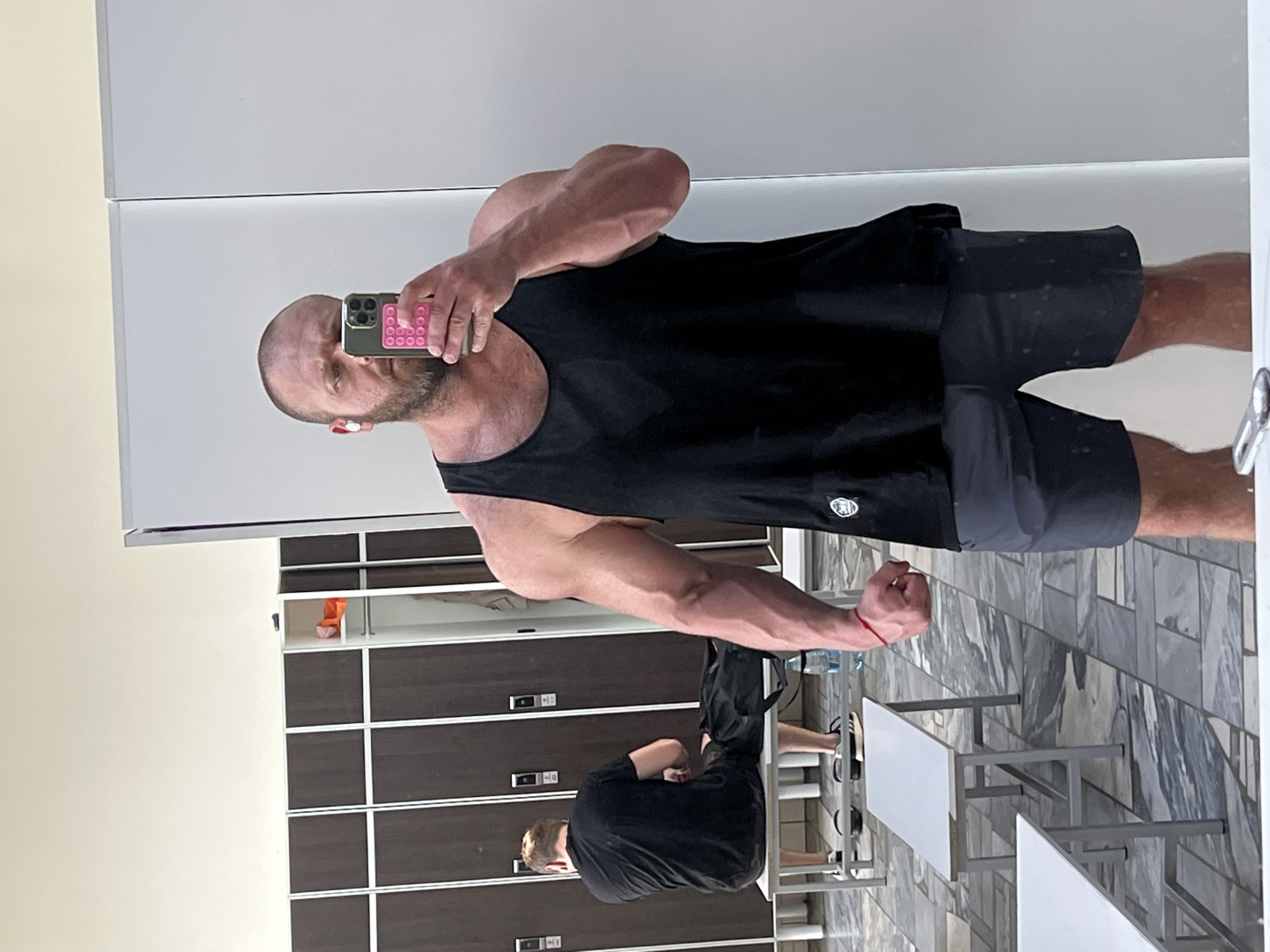 The image shows a man taking a mirror selfie in what appears to be a locker room or changing area. He is wearing a black tank top and grey shorts, and has a beard and bald head. He is holding a smartphone in his right hand, which has a pink pop socket attached to the back. In the background, another man is sitting on a bench, wearing a black t-shirt. There are rows of lockers on the left side of the image and benches in the foreground. The floor is tiled. A bottle of water is visible on the floor next to the bench. The overall impression is of a gym or sports facility.