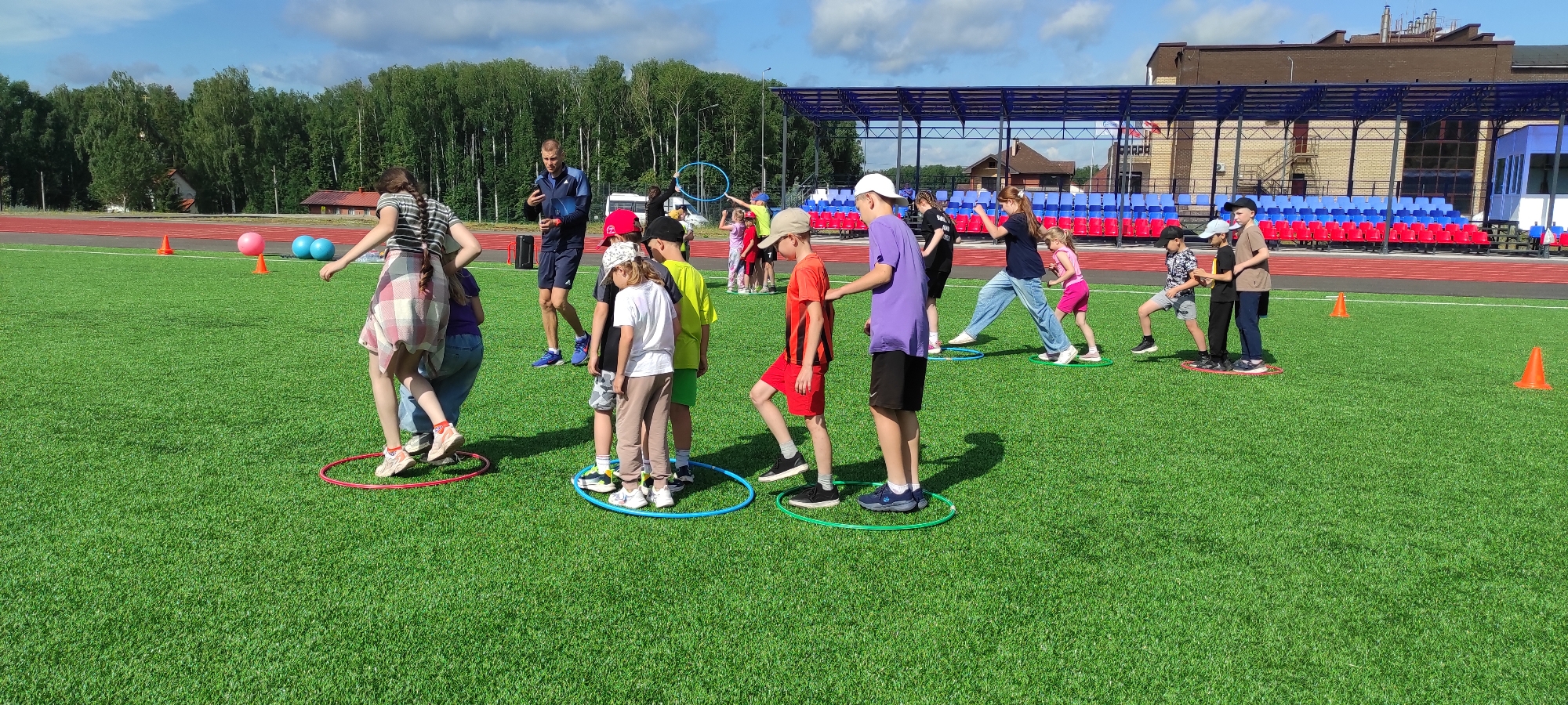 In Noviy, Russia, on a bright, sunny day, a group of children and an adult male are engaged in an outdoor activity on a vibrant green, artificial turf field, likely part of a sports complex or school grounds. The field is marked with white lines and has a running track adjacent to it. In the background, a tiered seating area with red and blue seats and a covered structure can be seen, suggesting a stadium or arena. Trees form a dense line behind the stadium.
The primary focus of the image is a group of children participating in a game involving hula hoops placed on the ground. A girl with braided hair, wearing a striped shirt and a checkered skirt, is stepping into a red hula hoop. Another girl in a purple shirt and jeans is behind her. To the right, a group of boys are also interacting with hula hoops. One boy in a purple t-shirt and black shorts is standing in a blue hoop. Another boy in an orange t-shirt and red shorts is stepping into a green hoop. Further to the right, more children are scattered across the field, some participating in the hoop activity and others seemingly observing or moving between stations. A man in athletic wear, possibly a coach or instructor, is standing near the center of the group, gesturing with his hand. Several orange cones are visible on the field, and a few pink and blue spherical objects, possibly balls, are situated in the background. The overall atmosphere appears to be one of active recreation and organized play.