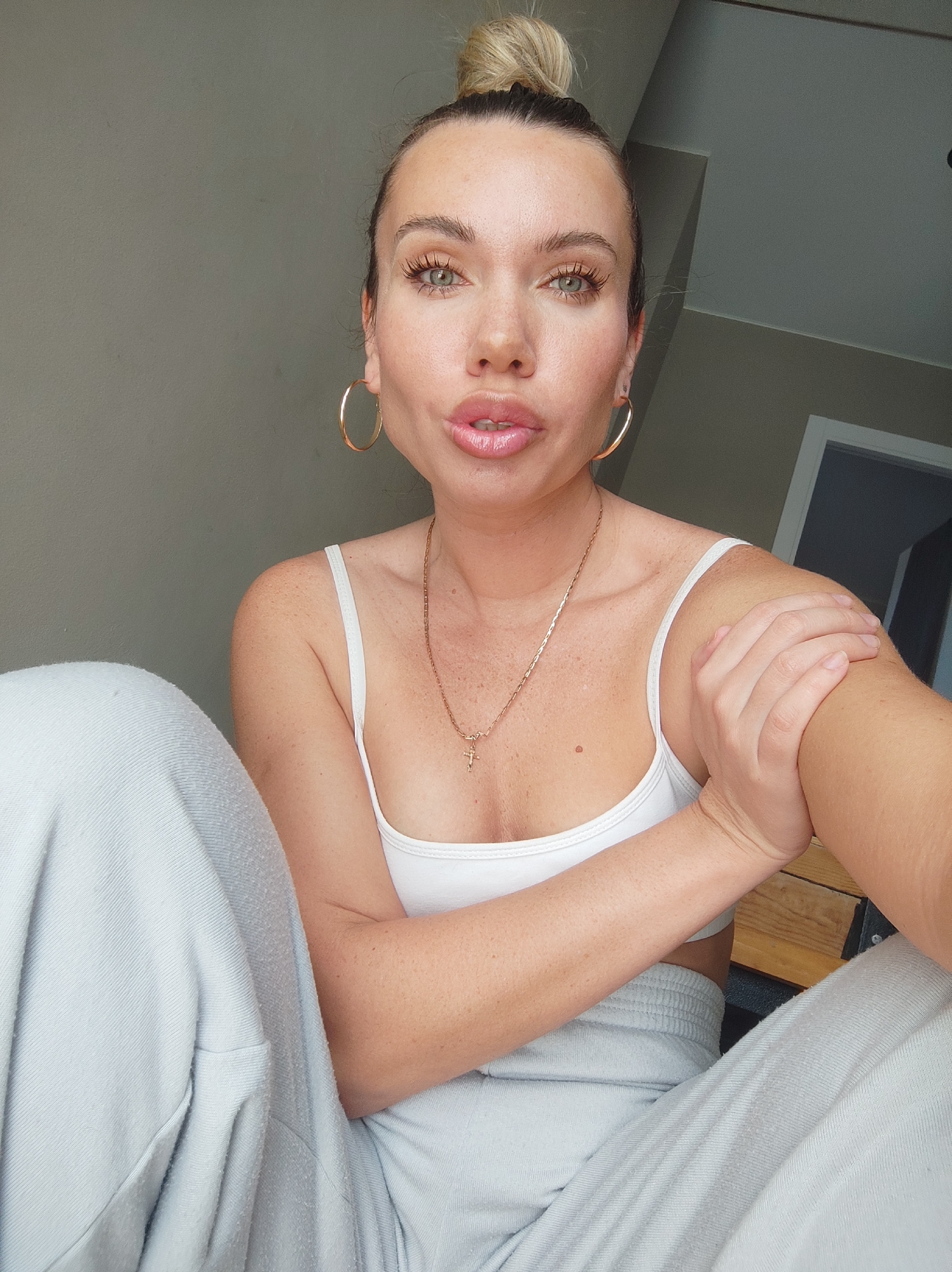 A woman is indoors, possibly in an apartment in Votkinsk, Russia. She has blonde hair in a bun, green eyes, and is wearing a white tank top and light grey sweatpants. She is also wearing large gold hoop earrings and a gold necklace with a cross pendant. She is sitting with her legs crossed and one arm holding the other. The background is a neutral grey wall, with a glimpse of a white door frame.
