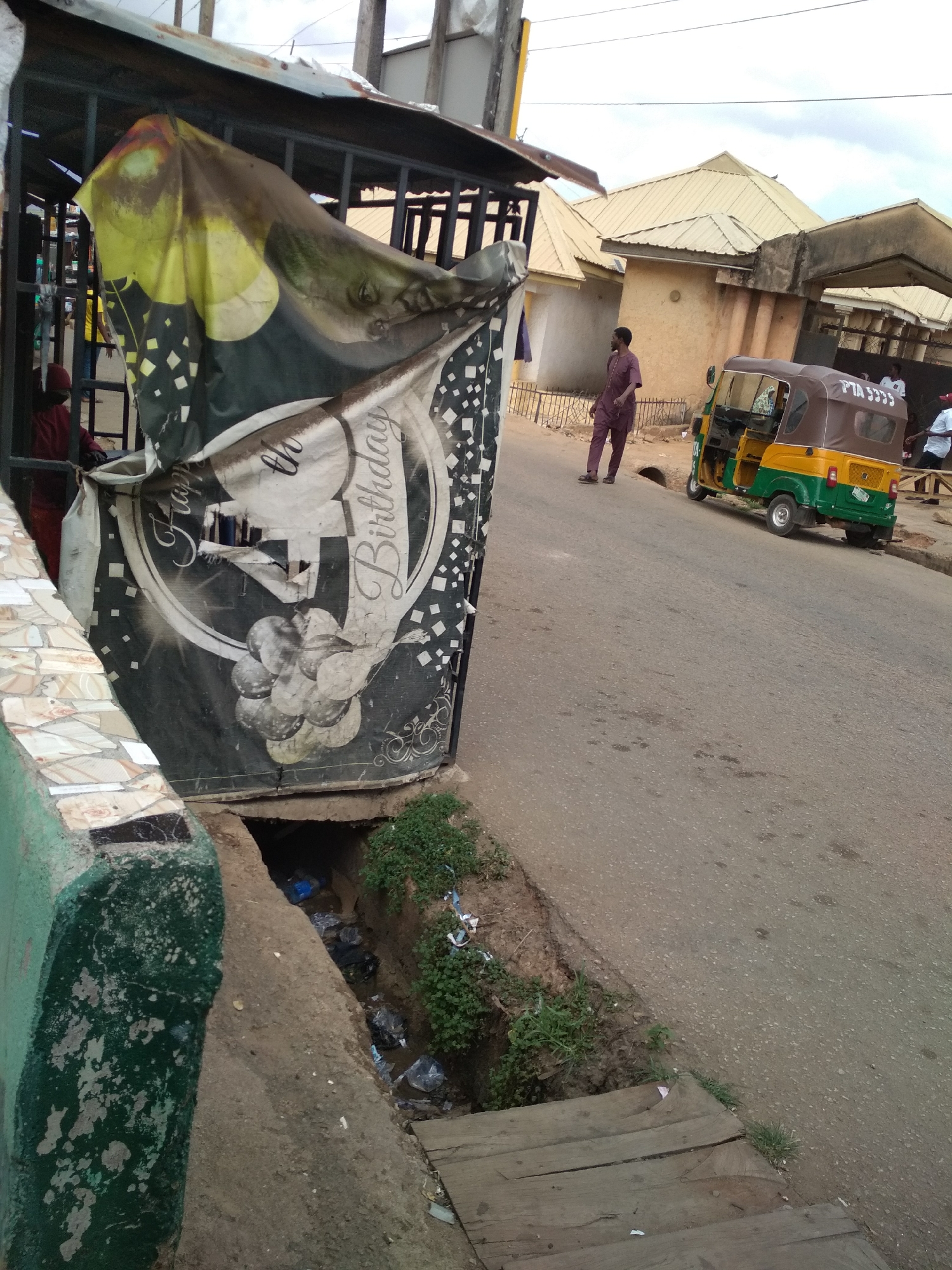 The image depicts a street scene in what appears to be a residential area. A dilapidated "Happy Birthday" sign hangs on a metal structure near a green, mosaic-tiled wall. A ditch filled with trash runs along the side of the road, covered by a wooden plank. A man in maroon attire walks on the road beside a green and yellow auto rickshaw, commonly known as a "Keke Napep," a common means of transport in many Nigerian cities. Other individuals can be seen in the background near a building. The scene suggests a location in a lower-income area, possibly Jos, Nigeria, inferred from the architectural style and the prevalence of Keke Napeps.