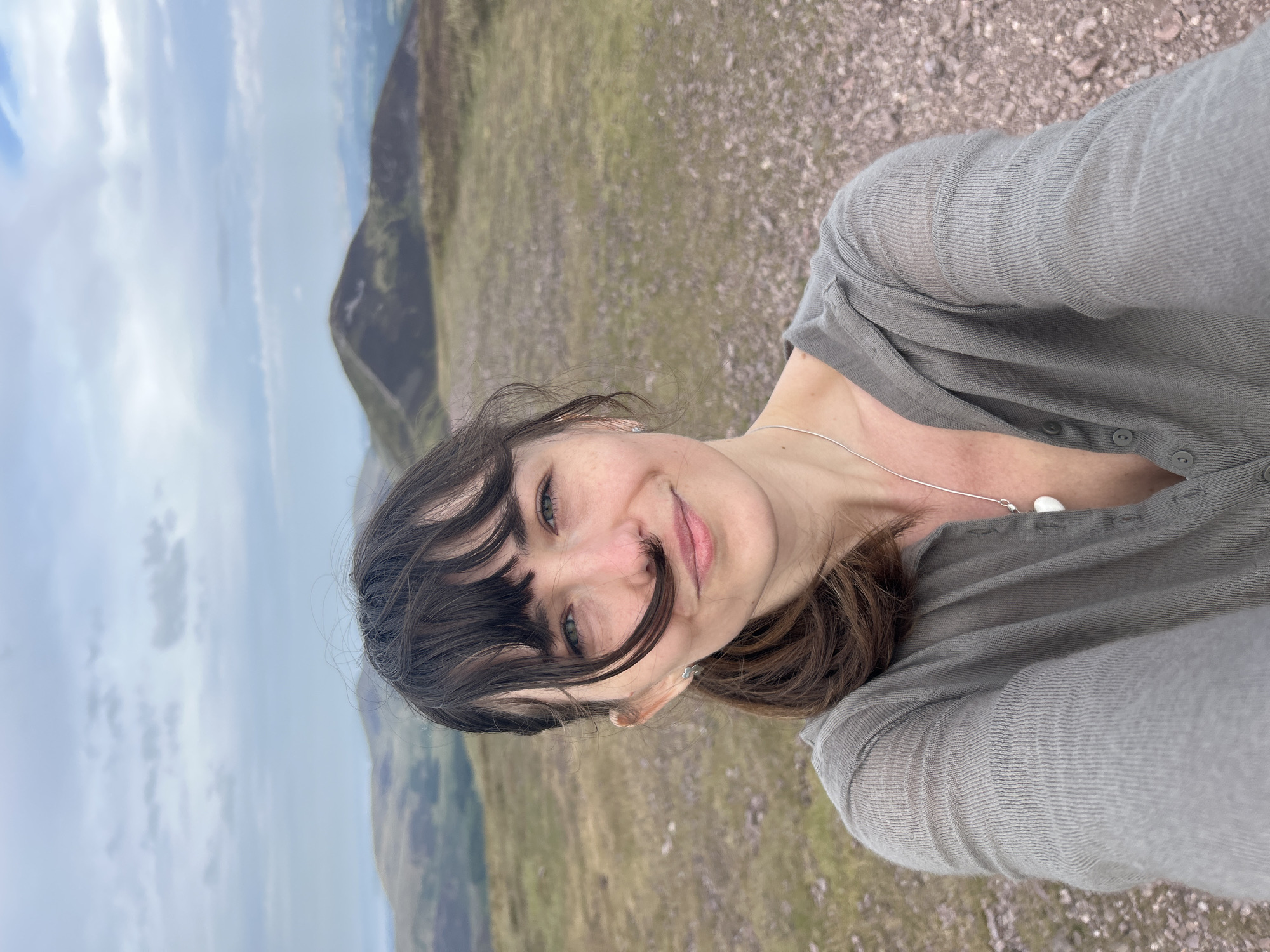 The image is a selfie taken outdoors on a hilly, barren landscape. The subject is a woman with dark hair and bangs, wearing a grey cardigan and a silver necklace with a heart-shaped pendant. She is smiling and looking towards the camera. Her hair is blowing across her face. In the background, there are rolling hills under a cloudy sky. The ground is covered in gravel and sparse vegetation. The overall scene suggests a hike or outdoor excursion in a rural location. The specific location in Penicuik, United Kingdom cannot be determined from the image alone.