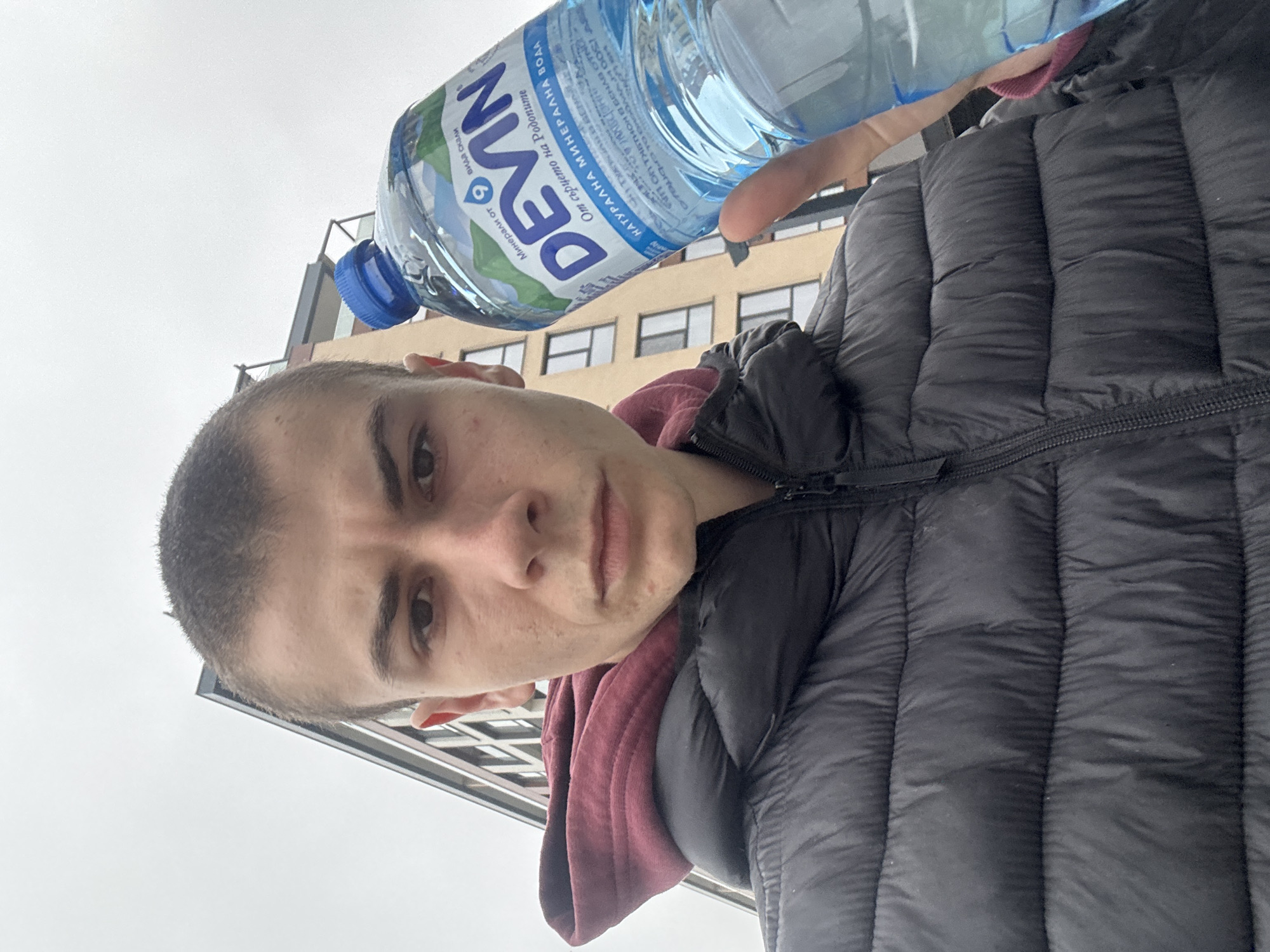 A person is holding a bottle of "Devin" mineral water in front of a multi-story building. The person has short hair and is wearing a black puffer jacket over a maroon hooded sweatshirt. The bottle label indicates it's from the Rhodope Mountains. The setting appears to be outdoors with overcast skies. The location might be Plovdiv, Bulgaria, due to the Bulgarian language on the bottle.