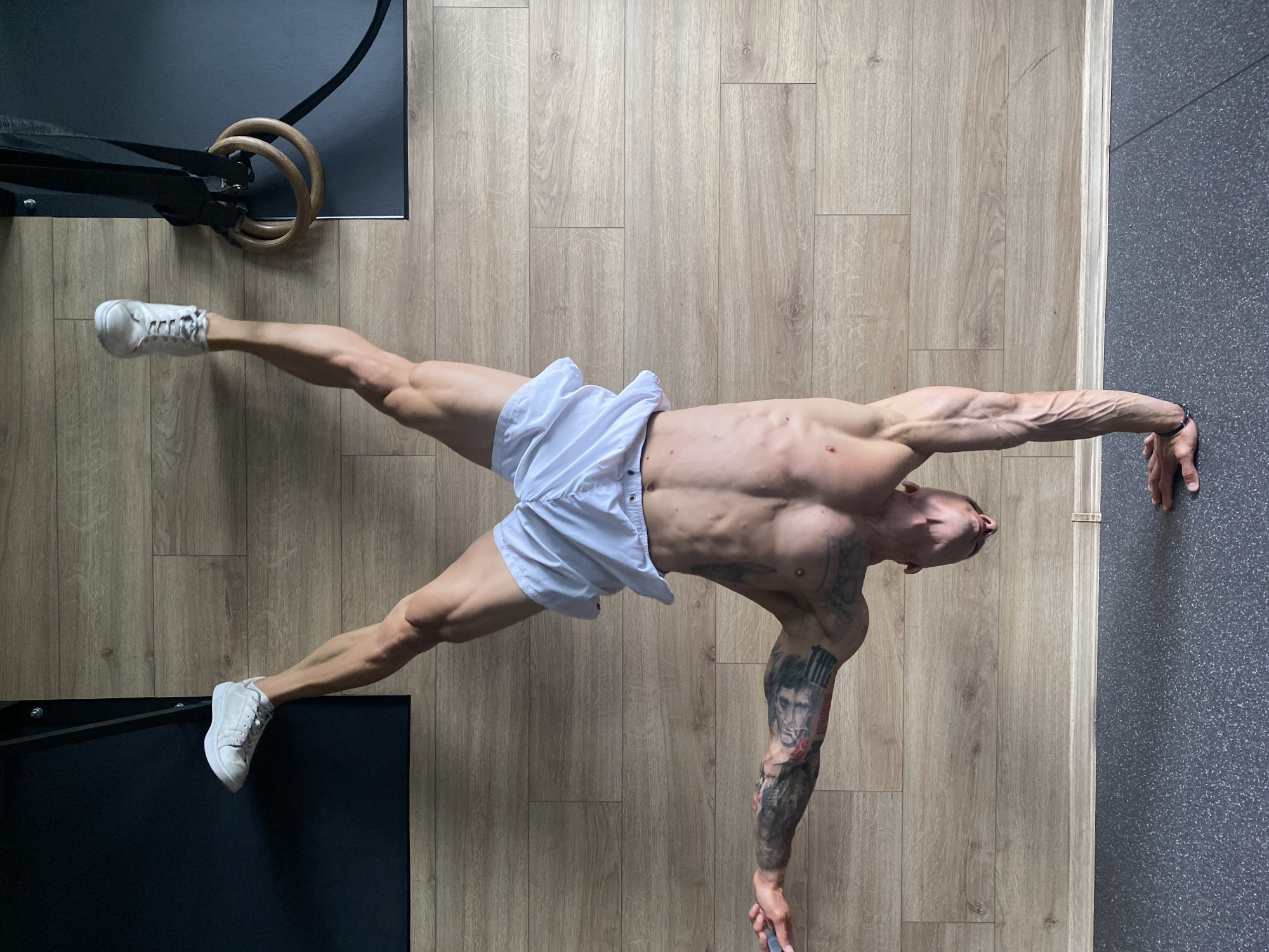 A man is performing a handstand in a gym. He is wearing white shorts and white sneakers. He has tattoos on his arms and torso. The gym has a wooden floor and a dark mat. There are gymnastic rings hanging from the ceiling. The image does not contain enough information to determine the location in the city of Pleven, Bulgaria.