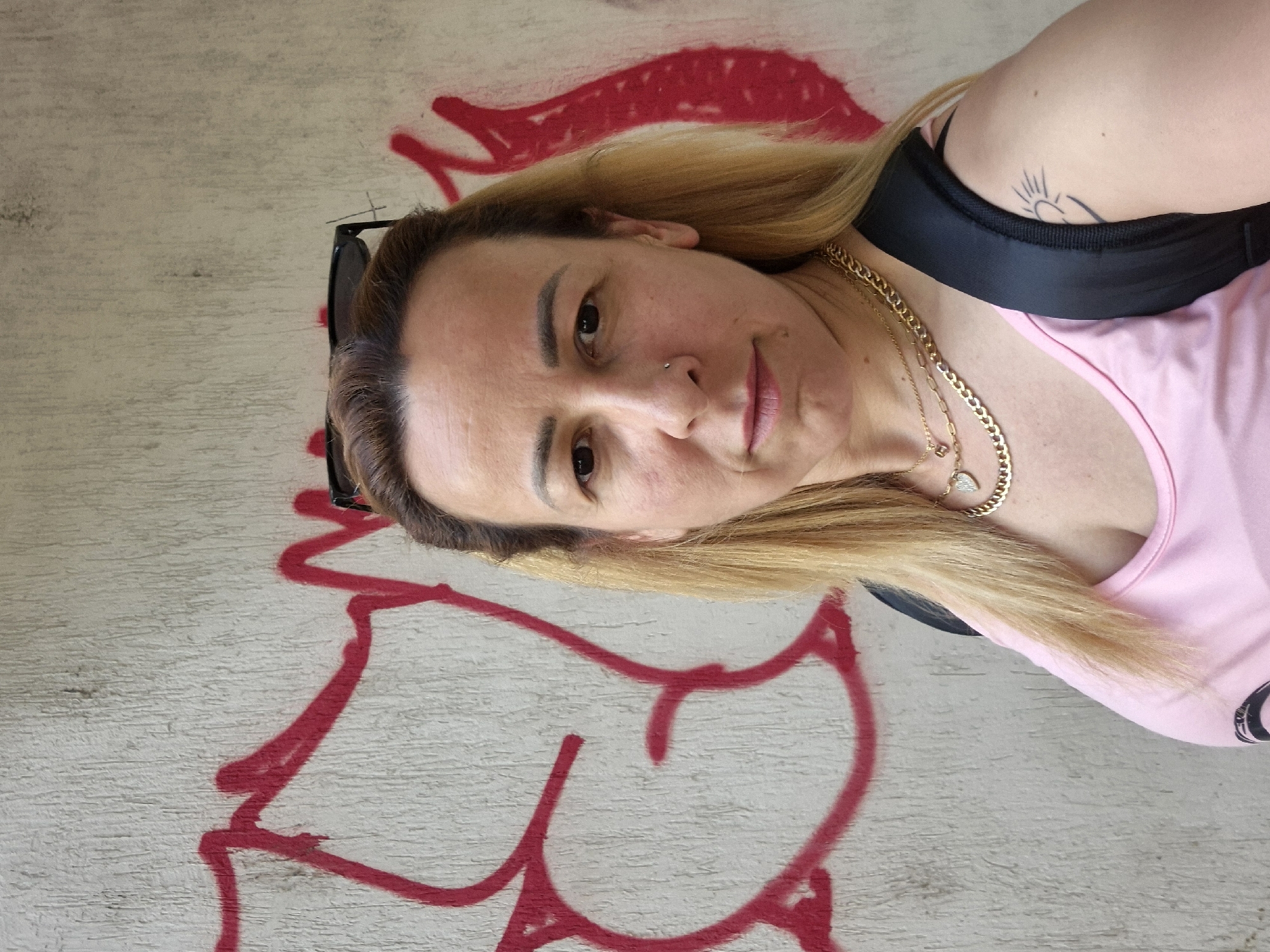 A medium shot captures a fair-skinned woman with shoulder-length blonde hair and a nose ring posing in front of a textured concrete wall adorned with red graffiti. She is wearing a pink tank top, a gold chain necklace with a heart pendant, and a black backpack strap is visible over her right shoulder. Sunglasses are perched on top of her head. Her left arm, visible in the lower right portion of the frame, has a tattoo of a sun. The graffiti on the wall features abstract red lines and shapes. The lighting suggests an outdoor setting, possibly during the day, with the sun casting minimal shadows.