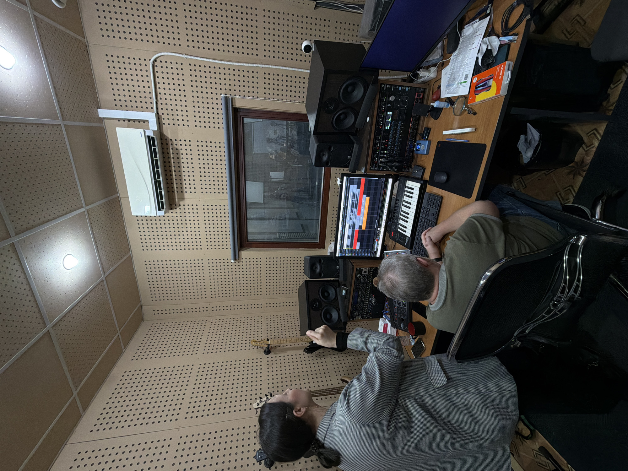 The image displays an overhead view of a music recording studio or control room located in Krasnoyarsk, Russia. The scene features two individuals actively engaged in a music production session.
The room's interior is characterized by light brown, perforated sound-absorbing panels covering both the walls and ceiling for acoustic treatment. A white split-system air conditioner, displaying "25" (likely the temperature), is mounted high on one wall. A window, likely soundproof, offers a view into an adjacent room, showing indistinct reflections. A small white security camera is positioned near the ceiling above the main workstation.
At a wooden desk, a person with gray hair and a beard, wearing a green short-sleeved top, is seated and intensely focused on two computer monitors. These screens display a Digital Audio Workstation (DAW) interface, showing multiple tracks, waveforms, and a piano roll, indicating audio editing or mixing. Various audio equipment surrounds the monitors, including large studio monitor speakers, smaller secondary monitors, and a rack-mounted audio interface adorned with numerous knobs and lights. A MIDI keyboard controller is placed in front of the screens. The desk surface is cluttered with a computer keyboard, mouse, a red mug, a smartphone, a magnifying glass, papers, cables, and an orange box labeled "ADATA".
Standing or leaning near the desk, a second person with dark hair tied back in a braid, wearing a gray and white checkered blazer, holds an electric guitar. Their posture suggests they are either playing, demonstrating a musical part, or preparing for a recording.
The overall scene conveys an atmosphere of focused collaboration and creative work within a professionally equipped audio production environment. The specific time of day is not discernible due to the indoor setting and artificial lighting.