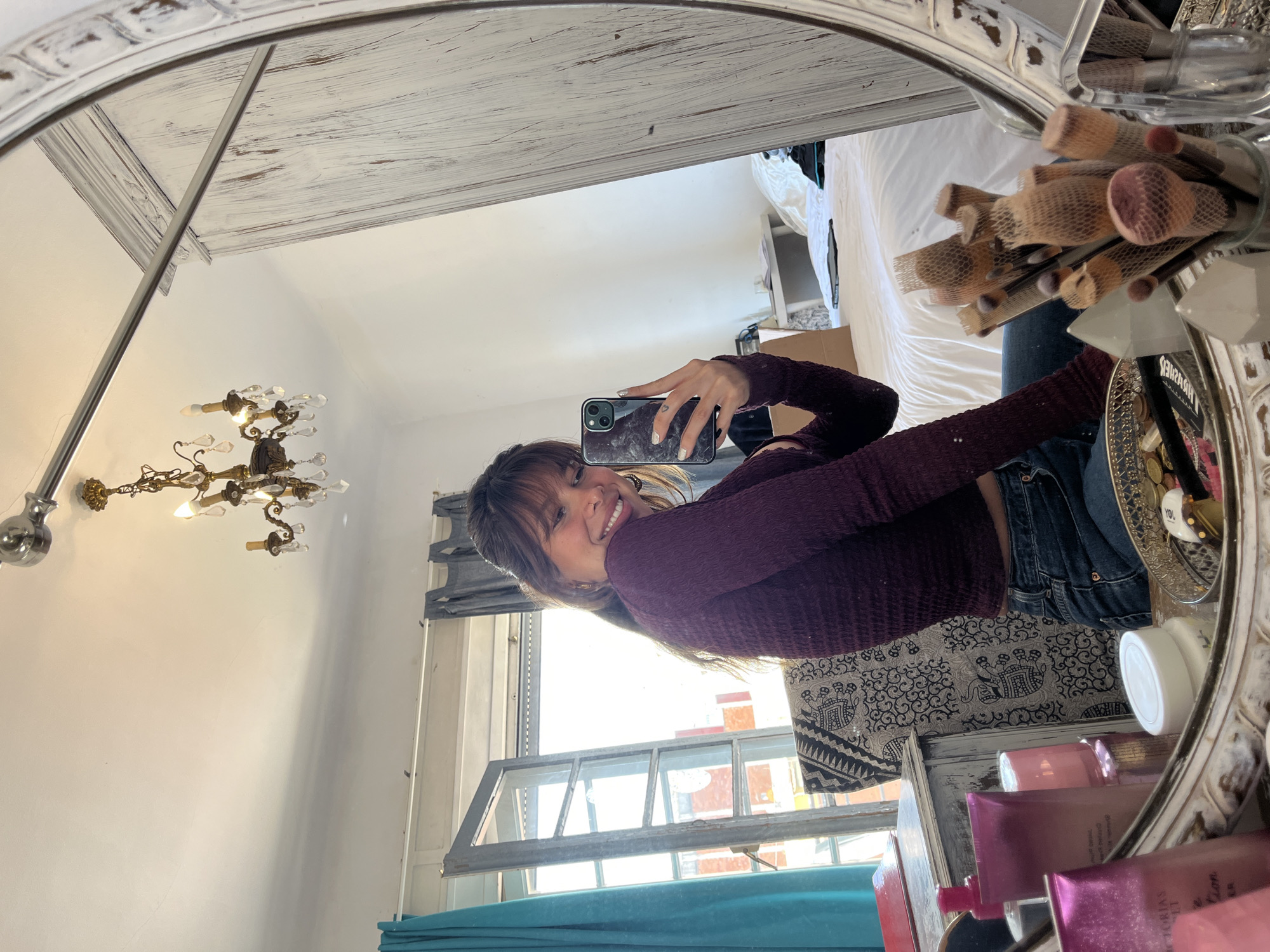 A young woman is reflected in an oval mirror, holding a smartphone to take a selfie. She's wearing a long-sleeved, dark purple shirt and blue jeans. Her face is visible, smiling toward the camera. Behind her in the mirror's reflection is a room interior, with a bed partially visible in the upper right, a window with blue curtains in the lower section, and a chandelier on the left wall. A variety of makeup brushes, beauty products, and decorative objects are clustered around the mirror's edge. The mirror appears to be placed on a vanity or table. There is no evidence to suggest this image was taken in Barcelona, Spain.