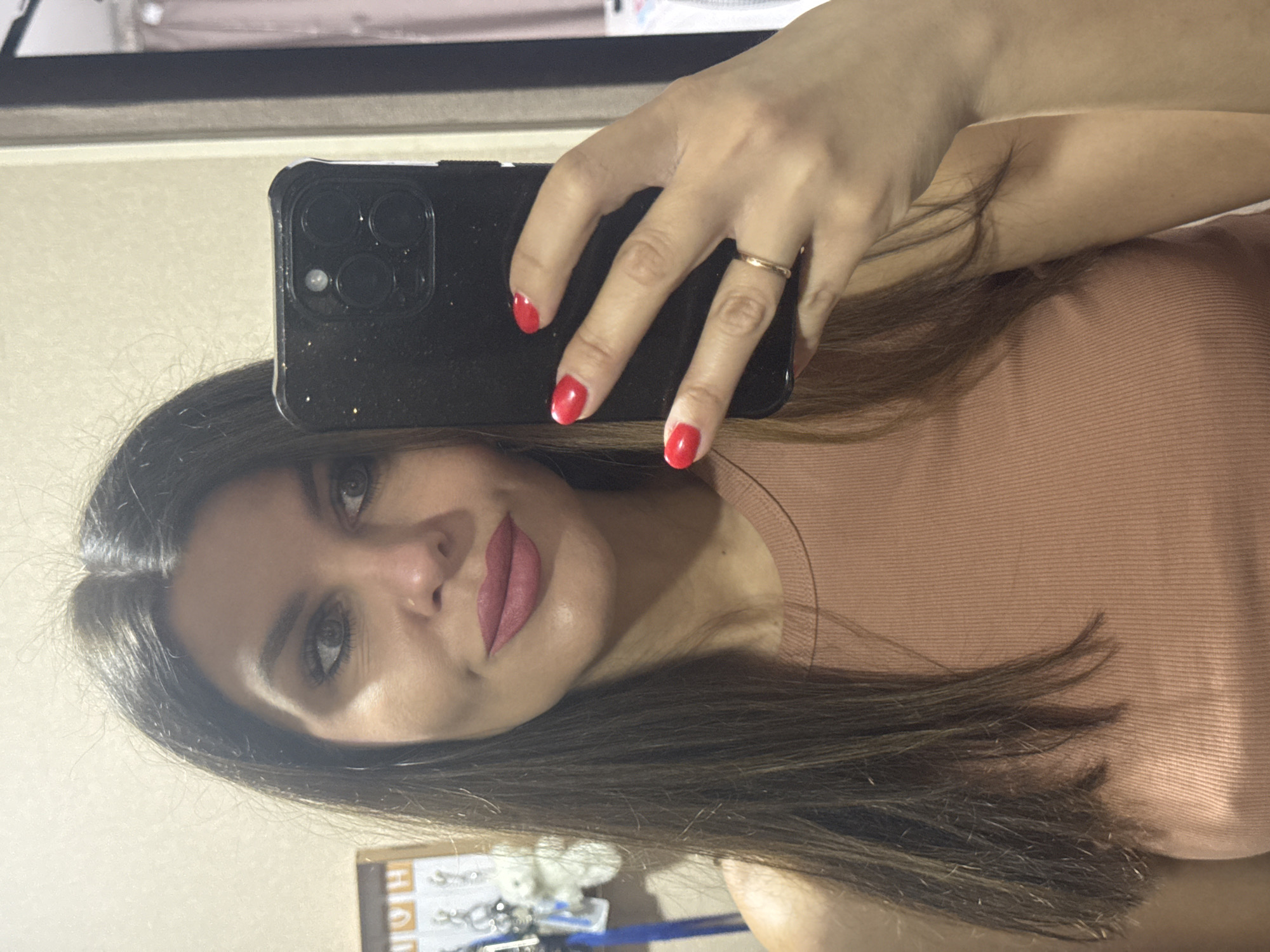 A woman is taking a selfie in a mirror. She has long, dark brown hair and is wearing a peach-colored top. Her nails are painted red, and she is wearing a ring on her left hand. In the background, a black cell phone is visible. The setting appears to be indoors, possibly a home or dressing room. There is no indication of the city of Chaikovsky, Russia in the image.