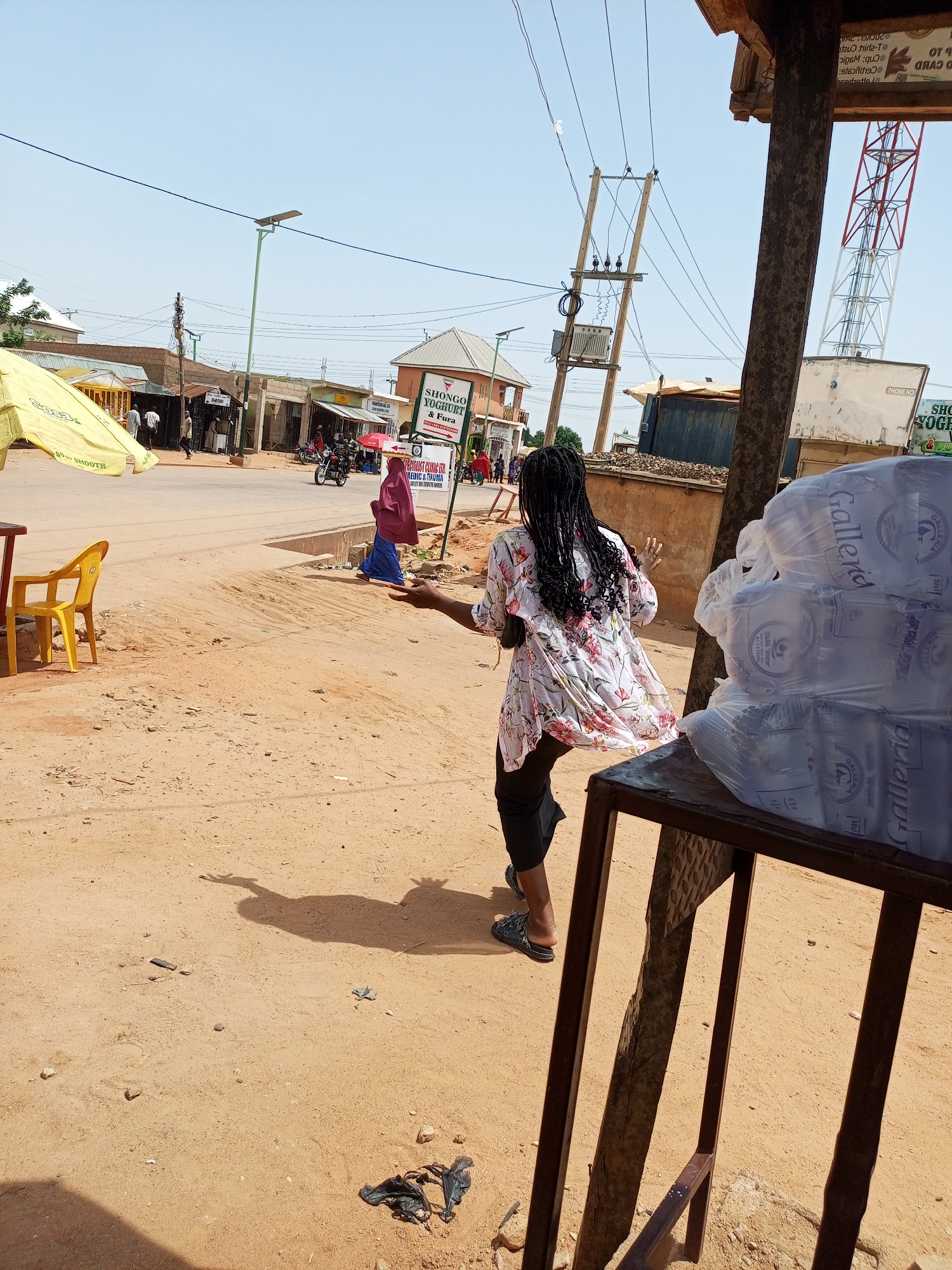 In Gombe, Nigeria, a young woman with long, dark braided hair walks away from the camera, her back to the viewer. She wears a floral-patterned blouse and dark trousers. The environment is an outdoor, sandy area next to a street with a few shops visible in the background. A yellow umbrella is set up on the left, and a yellow plastic chair sits nearby. A red and white telecommunications tower is visible in the distance. Several large bottles of water are stacked on a metal display rack in the foreground, partially obscuring the woman from the chest down. The ground is sandy and uneven, with some debris scattered about. The sky is clear and blue, indicating a sunny day.