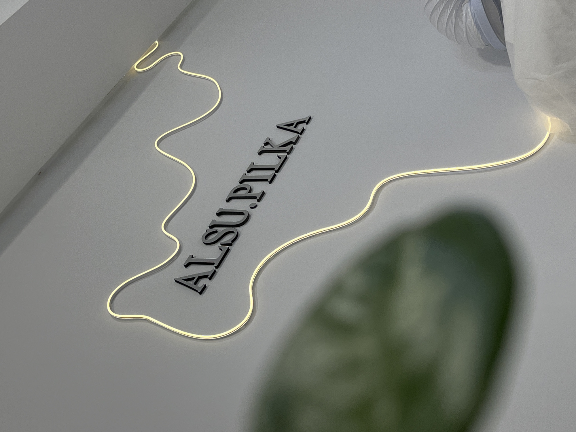 An interior scene, likely a commercial establishment in Neftekamsk, Russia, displays a white wall with a prominent mounted sign. The sign features dark, raised, serif-style lettering spelling "AL SU. PILKA." A thin, warm-white LED light strip in an irregular, wavy contour outlines and illuminates the text against the wall. A large, out-of-focus, dark green object, consistent with foliage, occupies the lower left portion of the foreground. In the lower right, a segment of a white, ribbed, flexible tube, possibly a ventilation or vacuum hose with a light blue band, is visible resting on a draped white fabric or sheet. No individuals or active interactions are present in the frame.