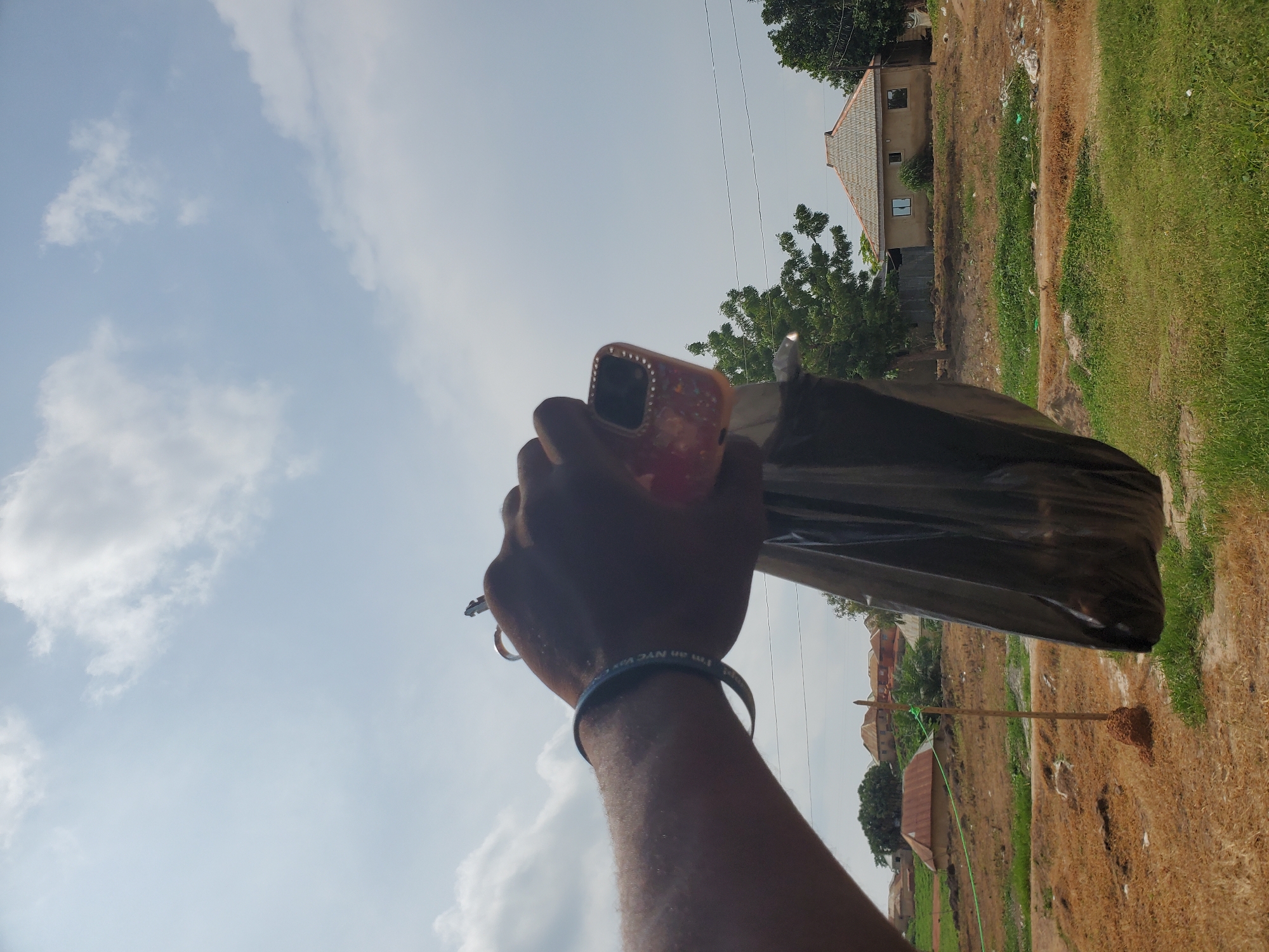 The image shows a person's hand holding a black plastic bag and a pink cell phone case against a backdrop of a cloudy sky and a rural landscape. The hand has a blue bracelet on the wrist, and two rings are visible on the fingers. The cell phone case is adorned with rhinestones. In the background, there are several houses with tiled roofs, trees, and dry vegetation. The ground appears to be uneven and scattered with some debris. The scene suggests an outdoor setting, possibly in a village or rural area. The location of Winters, United States of America, is not discernible from the image itself.