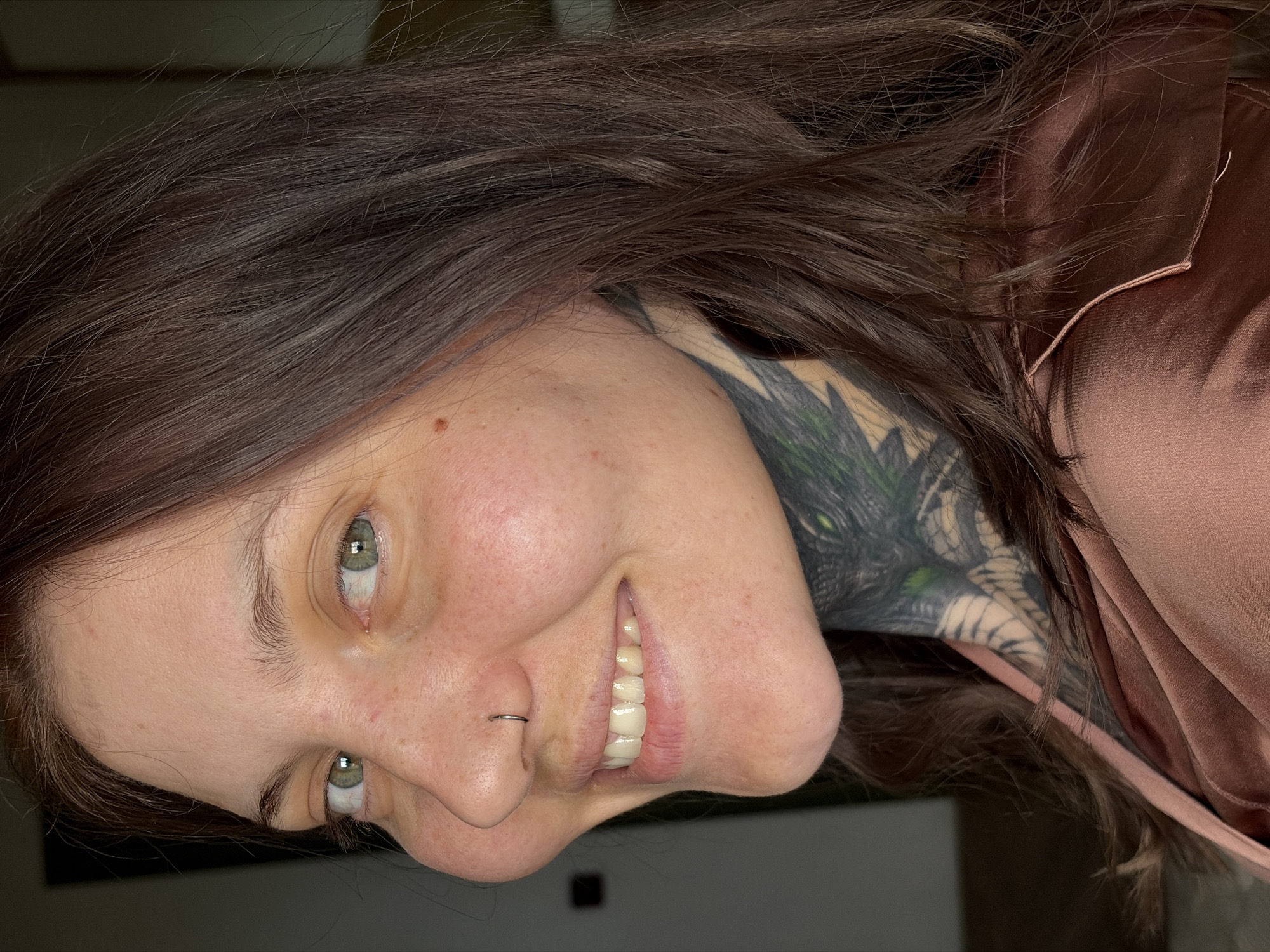 A female individual is depicted in a close-up, head-and-shoulders portrait, facing the viewer's left with her head turned to look directly at the camera. She has long, dark brown hair, and light green eyes. A small silver-colored hoop nose ring is present on her left nostril. She displays a wide smile, revealing her upper teeth. Her skin has a fair complexion, with visible pores and some minor blemishes or freckles on her face. A small brown mole is visible on her right cheek, near the hairline. A prominent, intricate tattoo covers the left side of her neck and extends downwards, featuring what appears to be a reptilian or dragon eye with green highlights and other patterns. She is wearing a light brown or rose gold-colored garment, possibly made of satin or silk, with a visible seam on the right shoulder. The background is dark and out of focus, providing no discernible environmental details. The exact location, including the city of Chaikovsky, Russia, is not ascertainable from the visual information provided in the image.