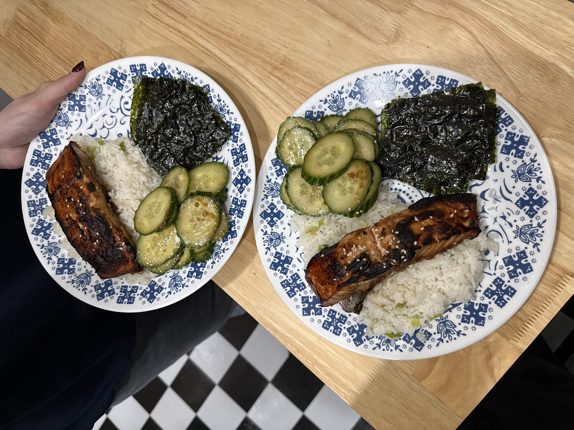 Here is a detailed description of the image:
The image shows two plates of food on a light brown wooden table. The plates are white with a blue floral pattern. Each plate contains a portion of rice, a piece of cooked salmon, a pile of sliced cucumbers, and a square of seaweed. The salmon is glazed and sprinkled with sesame seeds. The cucumbers appear to be coated in a light sauce. The rice has some green pieces. One plate is held by a person with dark red nail polish. The background includes a black and white checkered floor visible on the lower left side of the frame. The scene appears to be indoors.