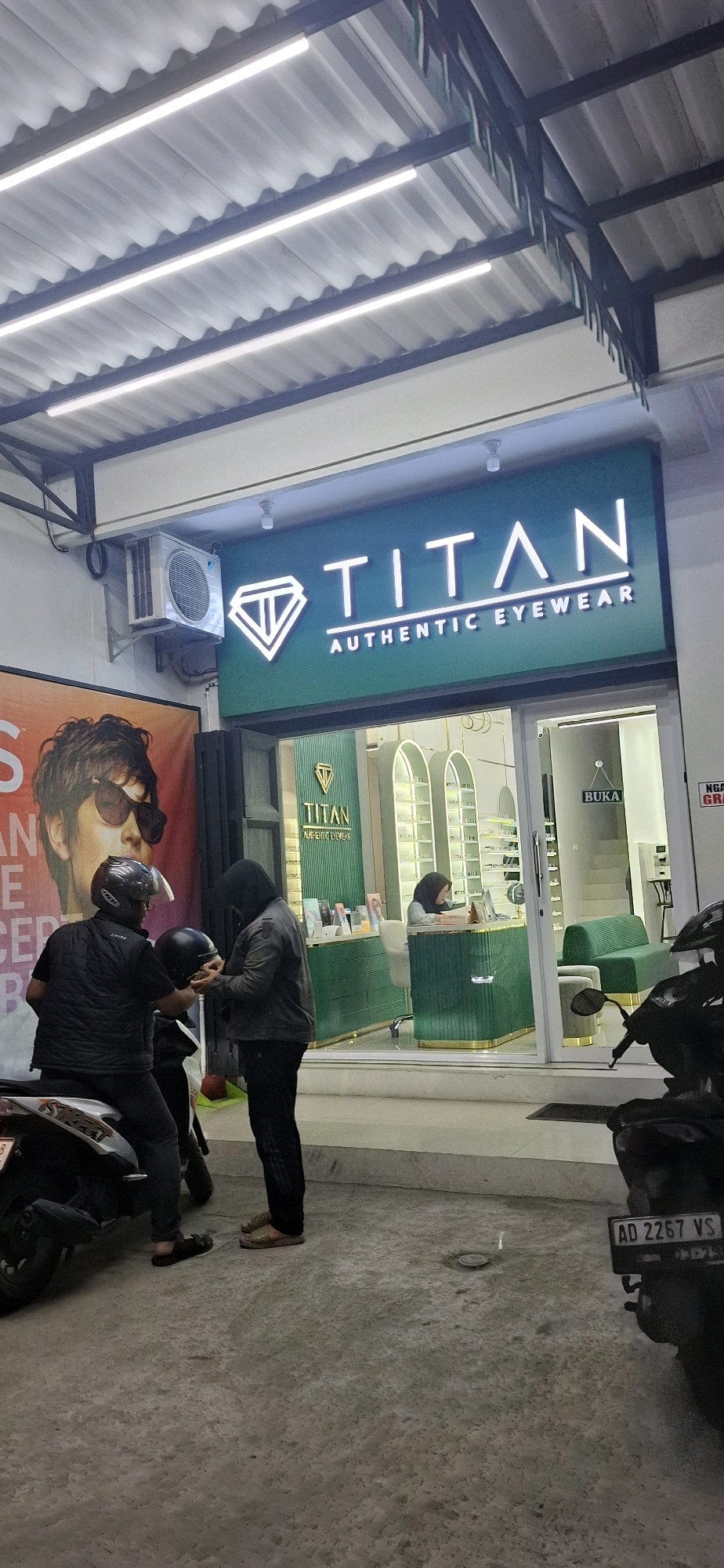 In Pabelan, Indonesia, this image captures the exterior of a store named "Titan Authentic Eyewear." The store's facade features a prominent, illuminated green sign with white lettering that reads "TITAN AUTHENTIC EYEWEAR" and a diamond-shaped logo. The entrance to the store is visible, with a "BUKA" (Open) sign on the glass door. Inside, a person is seen at a counter, and shelves displaying eyewear are visible.
Outside the store, two individuals are standing near a motorcycle. One person is seated on the motorcycle, wearing a black vest and helmet, and appears to be handing a black helmet to the other person who is standing and wearing a dark jacket and head covering. Another motorcycle is parked to the right. The ground is paved concrete, and an air conditioning unit is mounted on the wall above the store entrance. A large advertisement poster featuring a man wearing sunglasses is partially visible to the left of the store entrance. The overall scene suggests a commercial street view.