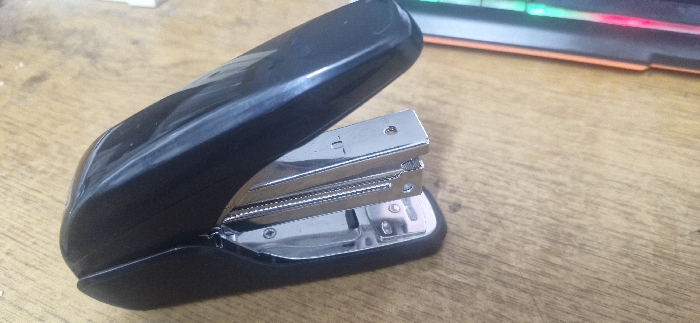 The scene, located in the city of Yantarniy, Russia, displays a black stapler positioned diagonally on a wooden surface. The stapler is in an open state, revealing its internal silver-colored metal mechanism, which includes a staple track and an impact plate. A "T-" marking is visible on the upper part of the metal mechanism. The stapler rests on a light brown wooden surface with visible grain patterns. In the upper right background, out of focus, a portion of a black object with illuminated green, red, and orange lights is discernible, consistent with a backlit keyboard. A thin orange stripe is also visible below this object in the background. No people are present.