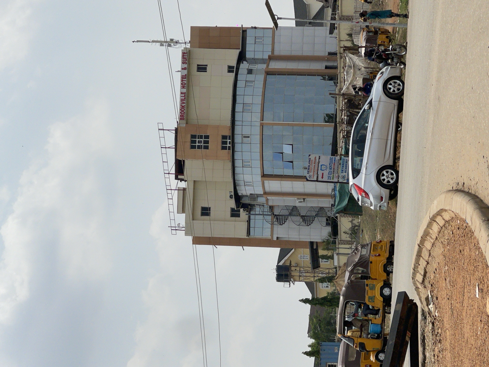 This media file depicts a street scene in Abuja, Nigeria, under an overcast sky. The central subject is a multi-story building identified by a prominent sign as "BROOKVILLE HOTEL & SUITE." The building features a contemporary design with large sections of reflective glass windows contrasting with cream and light brown facade panels. External staircases or maintenance structures are visible on the building's side.
In the foreground, a paved road runs parallel to a dirt or sandy shoulder, marked by a concrete curb. A white sedan is parked on the side of the road, facing left. Further down the street, several yellow tricycles, commonly known as Keke Napep, are visible, some with occupants. A few individuals are present near the vehicles and in the vicinity of the hotel building. Overhead power lines span across the upper portion of the image. The sky is uniformly light gray, indicating an overcast or hazy daytime.
Visible text includes:
* "BROOKVILLE HOTEL & SUITE" (on the main building)
* "GEMSLAND"
* "Inkogema Ahu" (partially visible under Gemsland)
* "ZOE KIDS MONTESSORI"
* "TEL 08069660999"
* "SKYKREST ACADEMY"
* "AREST"
* "YYA"