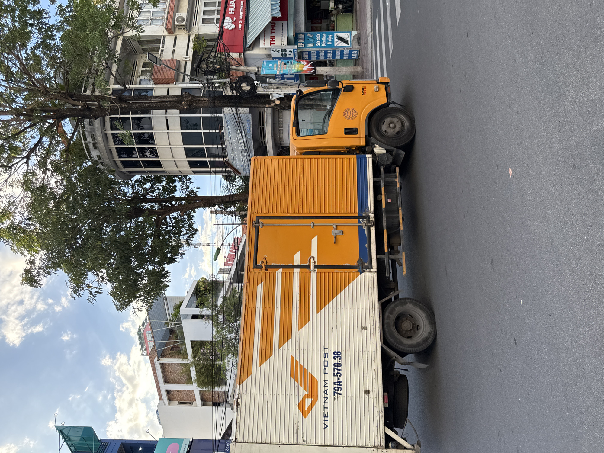 A bright orange and white box truck, identified as "VIETNAM POST" with license plate "79A-570.38", is parked on the right side of a paved city street in Nha Trang, Vietnam. The truck is positioned near a pedestrian crosswalk, indicated by white stripes on the road. In the background, several buildings are visible, including a modern white building with multi-level balconies and greenery, and a building with a distinctive curved, glass-fronted facade. Trees with full green foliage line the street, partially obscuring the buildings. The sky is partly cloudy with patches of blue. Various signs and utility poles are present along the street. The image appears to be taken during daylight.
