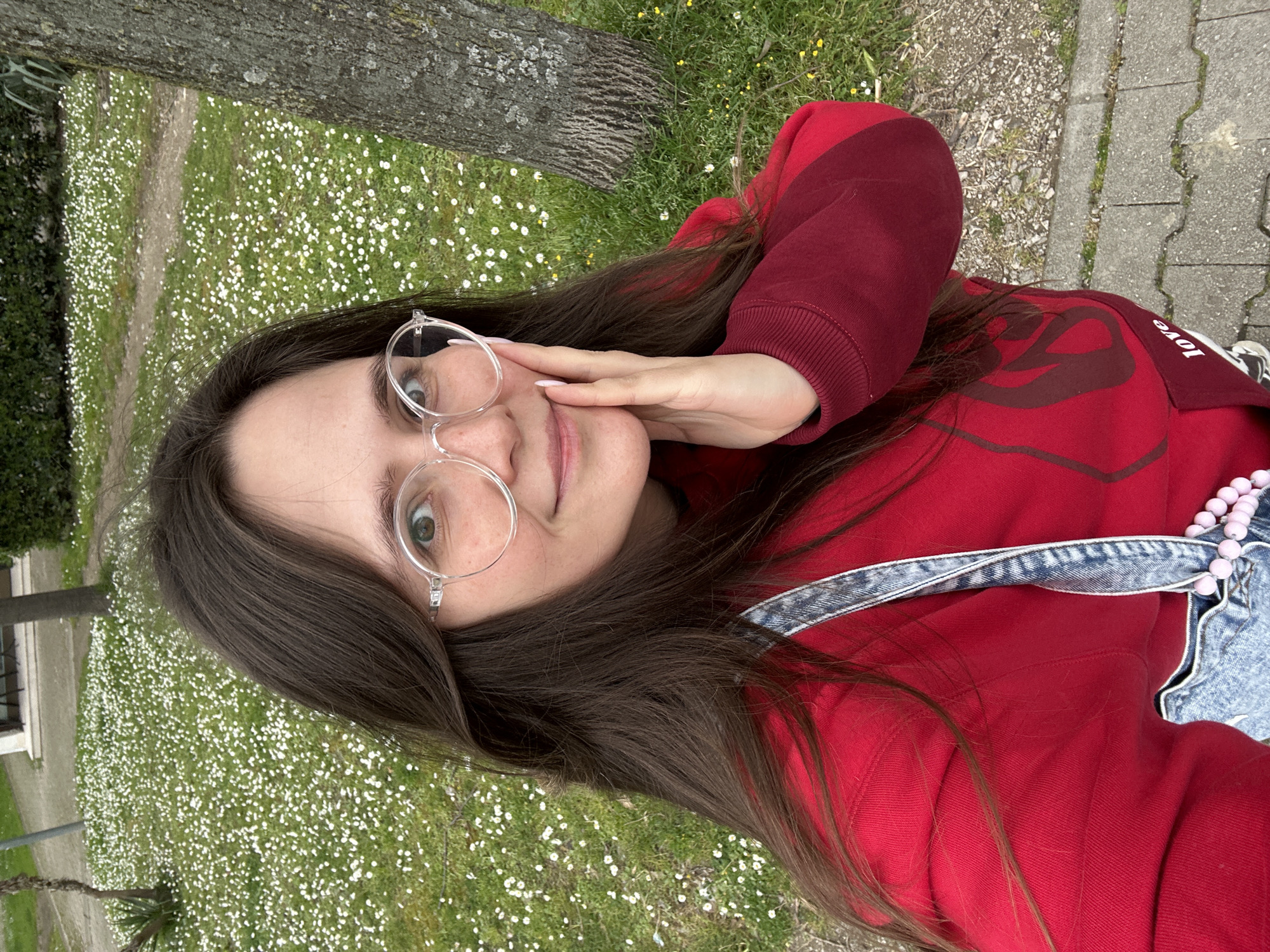 A young woman is posing for a selfie in an outdoor setting. She has long brown hair, light skin, and is wearing round, clear-framed glasses. She is wearing a red sweatshirt with darker red accents and a denim jacket with pink beads. She is touching her face with her hand. The background consists of green grass dotted with small white flowers. A tree trunk is visible on the left side of the frame, and a paved walkway is seen on the right. The location appears to be a park or green area, possibly within a residential neighborhood of Šušanj, Montenegro, based on the vegetation and surrounding urban elements.