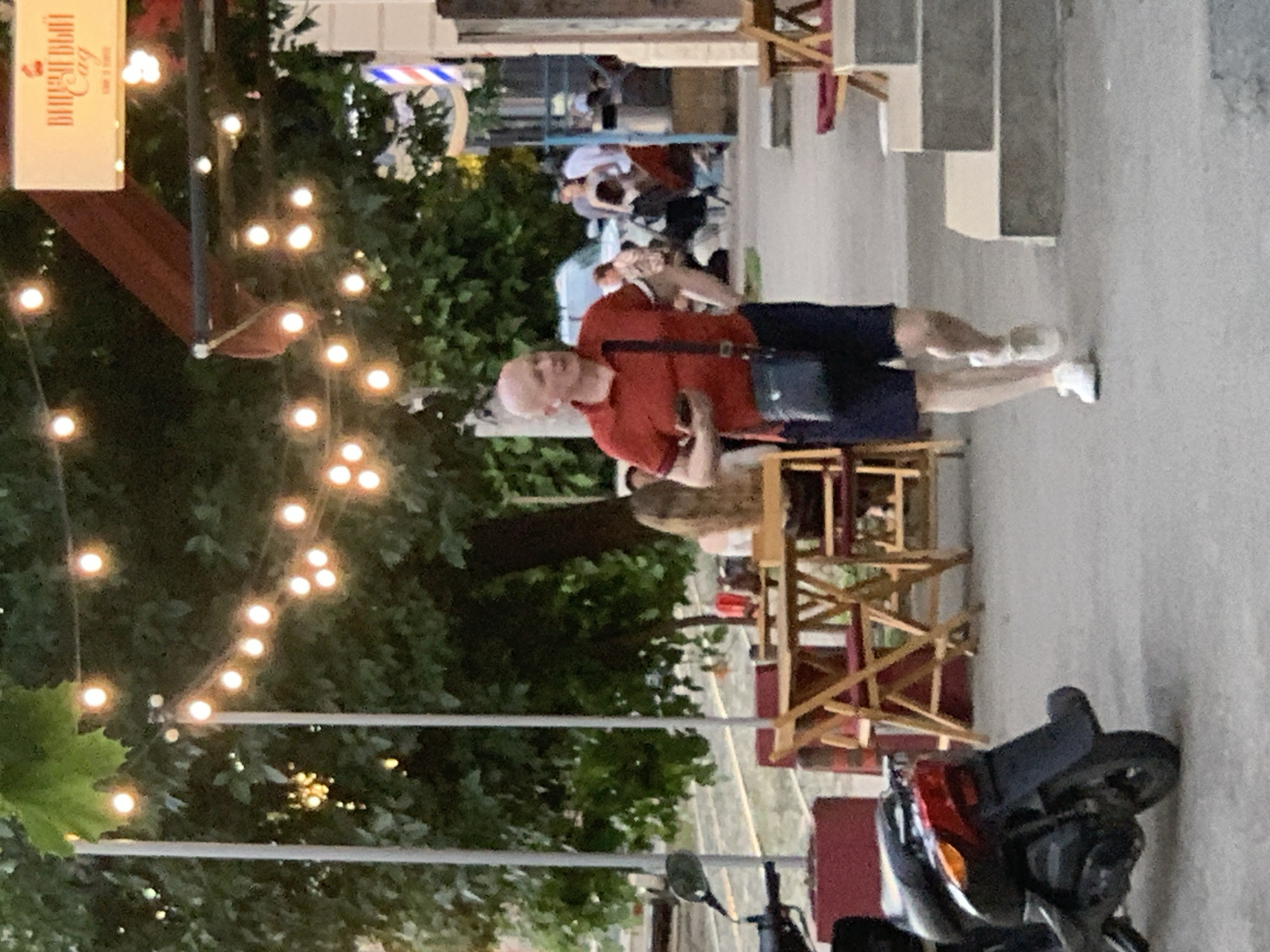 In an outdoor urban setting, a bald Caucasian man wearing a red polo shirt and dark shorts walks towards the viewer, holding a smartphone in his left hand and a black shoulder bag across his chest. He is wearing white sneakers. In the background, string lights illuminate the scene, and trees and foliage create a natural backdrop. Several tables and chairs are set up outside, suggesting a cafe or restaurant. A scooter is parked on the left side of the image. Other people are visible in the distance, seated at tables. The environment appears to be a pedestrian walkway or sidewalk.