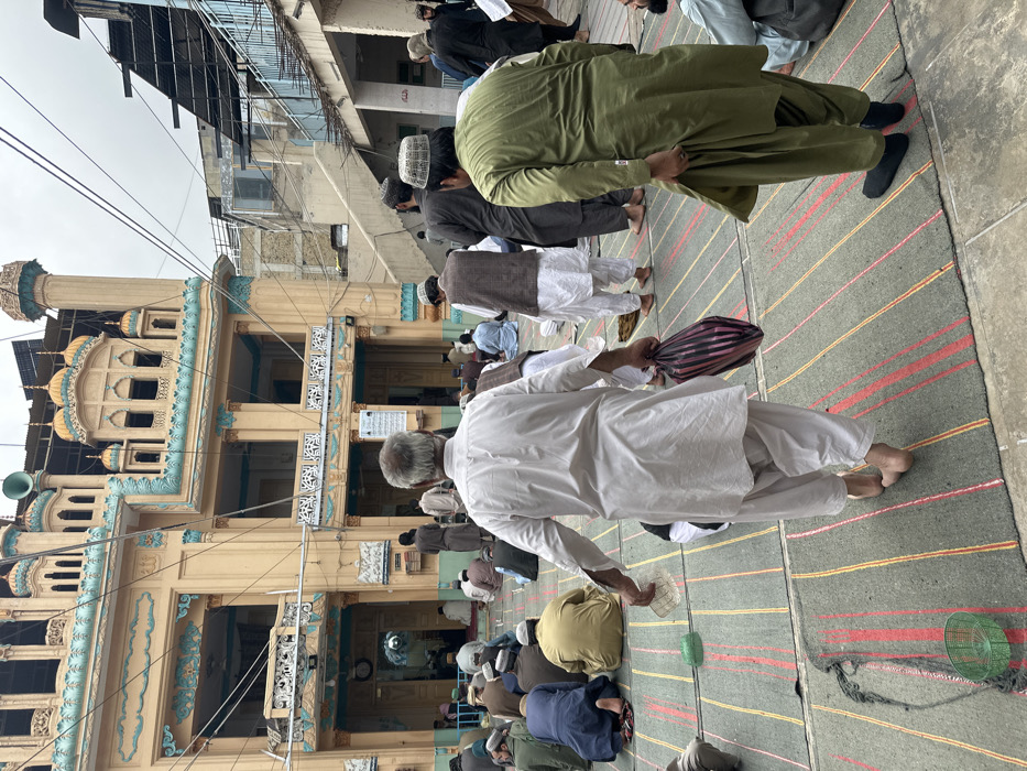 This image depicts an outdoor prayer area located in Quetta City Tehsil, Pakistan, under an overcast sky. The foreground and midground are covered with numerous rectangular, grey prayer mats featuring red and yellow stripes.
Approximately 30-40 adult males are present. Most individuals are seated or kneeling on the prayer mats, facing away from the camera, engaged in what appears to be communal prayer. They are predominantly dressed in traditional shalwar kameez attire; many wear various caps (kufi).
In the immediate foreground, two men are walking. An older man with grey hair, seen from behind, wears a white shalwar kameez and is barefoot. He holds a striped bag in his left hand and a clear plastic object in his right hand. To his right, another man, also seen from behind, wears an olive-green shalwar kameez and a white kufi cap, adjusting his garment with his right hand and wearing dark footwear or socks.
The prominent background structure is a multi-story mosque with a light beige facade, elaborate turquoise architectural trim, and multiple small domes, some gold-colored. Several minaret-like structures and a loudspeaker are visible on the roofline. Solar panels are also mounted on the mosque roof. Adjacent to the mosque on the right, less ornate buildings with visible staircases, railings, and exposed construction materials are present. Overhead electrical or communication wires crisscross the scene. Small green plastic baskets are visible on the prayer mats in the foreground.