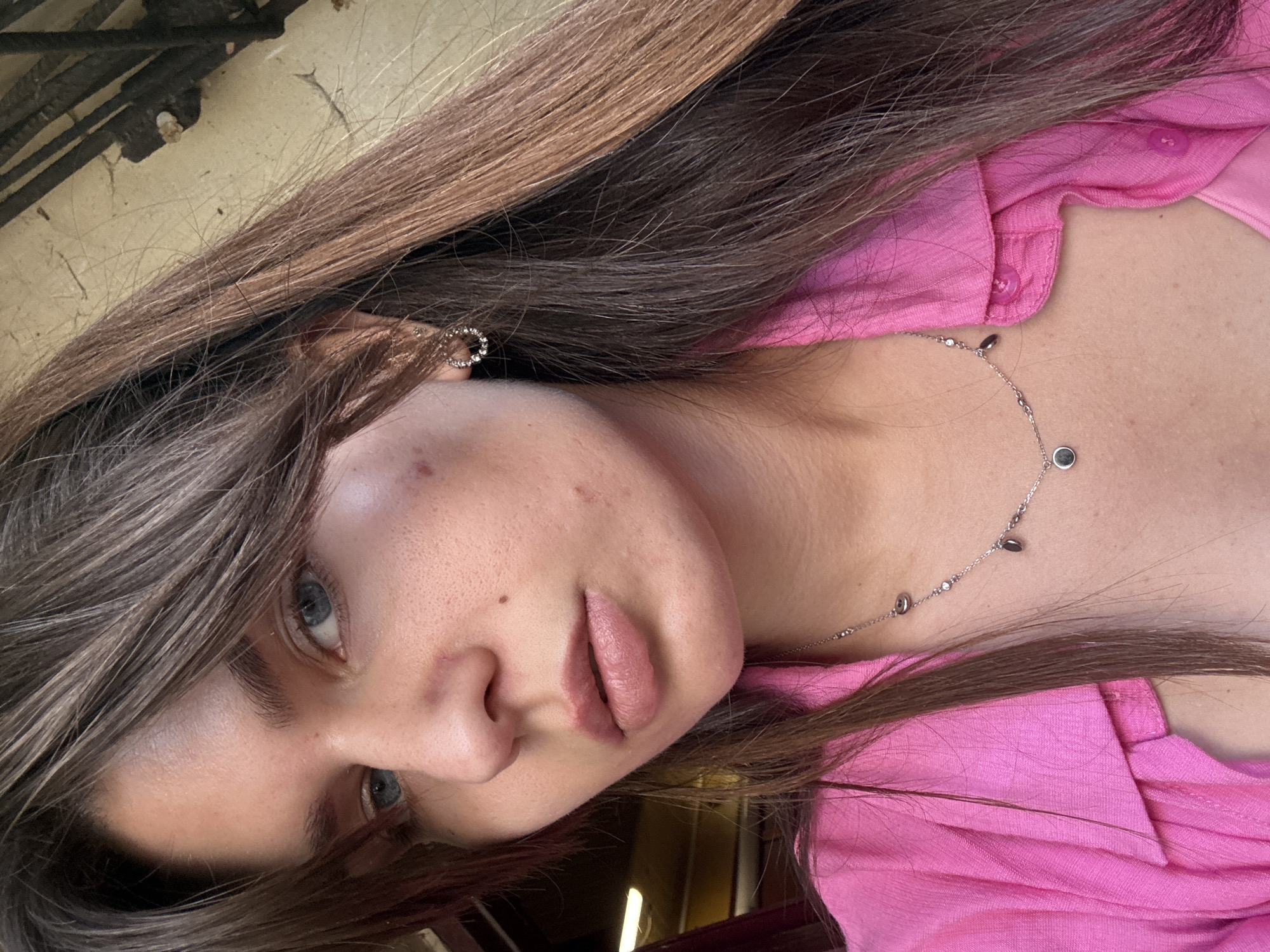 A woman with long brown hair and blue eyes is shown from the chest up. She is wearing a pink collared shirt with buttons. A delicate silver necklace with small charms hangs around her neck. She has a small hoop earring with rhinestones in her left ear. The background is a light-colored, textured wall.