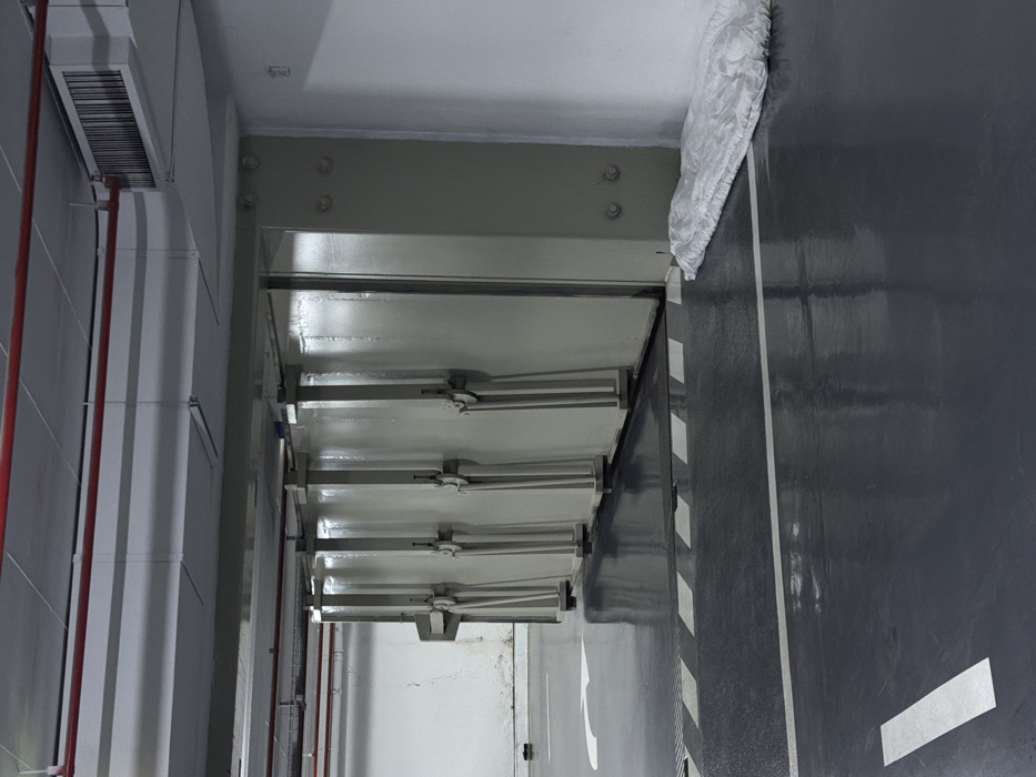 The scene depicts an interior view of an underground facility, likely a reinforced parking garage or shelter, situated in Tel-Aviv, Israel. A prominent feature is a row of four substantial, light-grey blast doors or sliding panels, each equipped with a visible manual locking lever mechanism, positioned along the right side of the frame. The floor is a dark grey, reflective surface marked with white painted lines, including a directional arrow on the left, a continuous solid line, and a striped pedestrian crossing pattern directly in front of the panels. A dark, textured mat abuts the base of these panels. The walls are light grey, exhibiting some discoloration at their lower sections on the left. The ceiling structure is light grey concrete, traversed by exposed red piping, likely for fire suppression, and grey electrical conduits. A rectangular ventilation grille is affixed to the upper right ceiling area. In the lower right corner, a crumpled white fibrous material, consistent with a tarp or large piece of fabric, lies against the wall. No individuals are present in the environment.