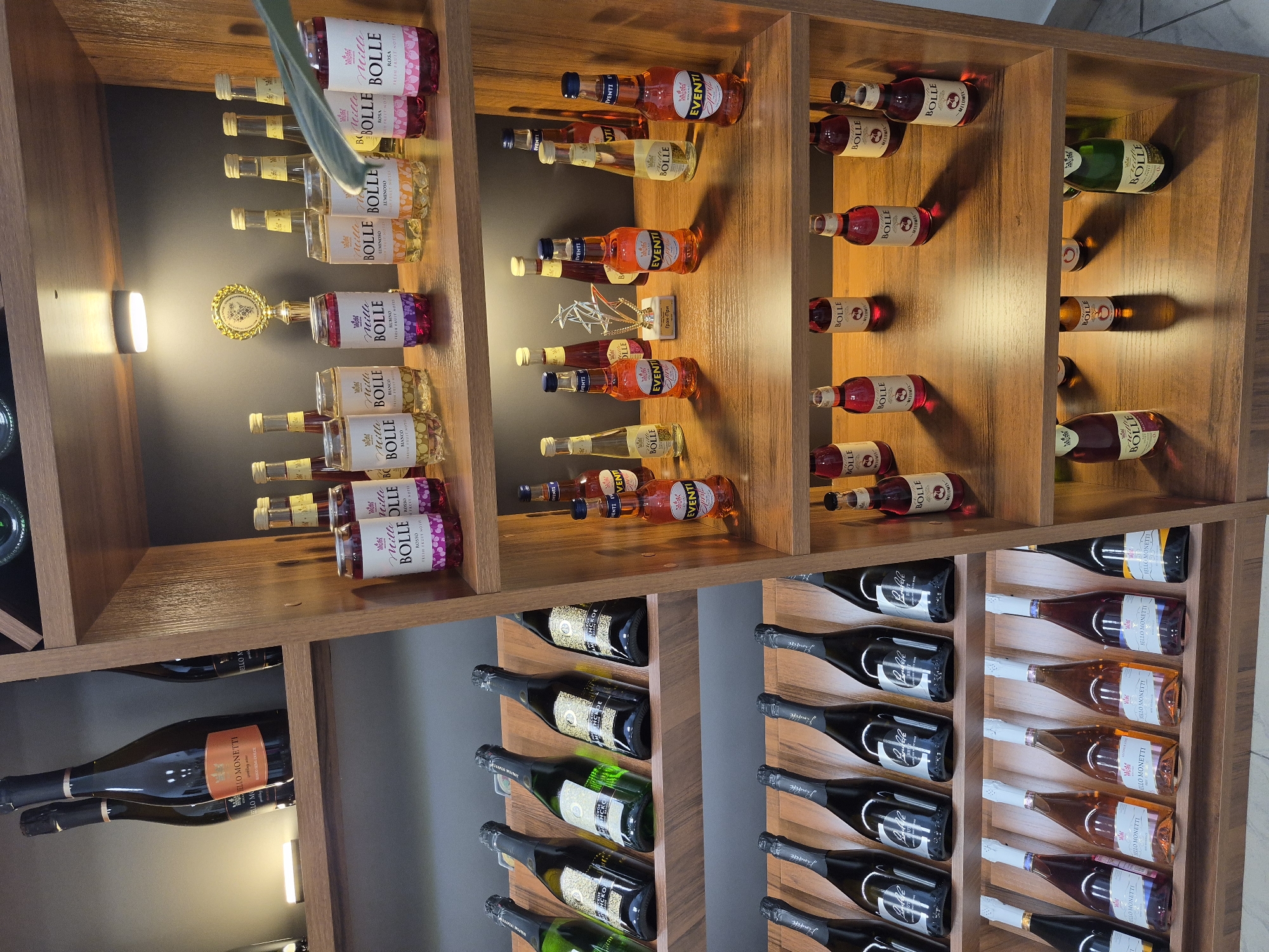 The image shows a display of bottled beverages, primarily wine and sparkling wine, arranged on wooden shelves in a retail setting. The setting appears to be a wine shop or a section of a larger store dedicated to alcohol sales. The lighting is warm and accentuates the products. On the top shelf, there are several bottles of sparkling wine with dark labels and gold lettering. Below that, there are two rows of sparkling wine bottles with various colored labels, some in green, black, and gold. The bottom shelf features a collection of smaller, colorful bottles, possibly aperitifs or flavored wines, with labels such as "BOLLE" and "EVENTI." A trophy is visible on one of the shelves, suggesting a display that might also be associated with awards or promotions. The overall impression is one of an organized and well-stocked retail environment for alcoholic beverages. The image does not contain any information about the city of Ufa, Russia.