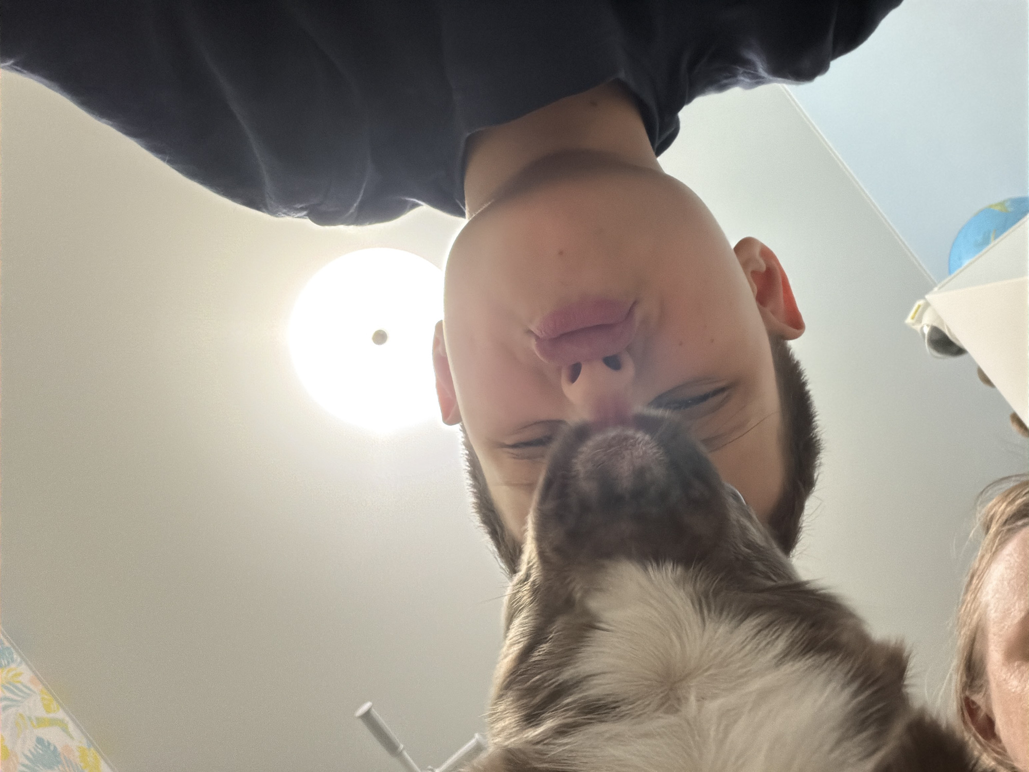 A young boy with pursed lips is close to a dog with white and brown fur. The boy's face is mostly in focus, while the dog's head is partially obscured, and some of the woman's face on the right side. A light fixture is in the background. The location is indiscernible from the image, and therefore cannot be determined to be Votkinsk, Russia.